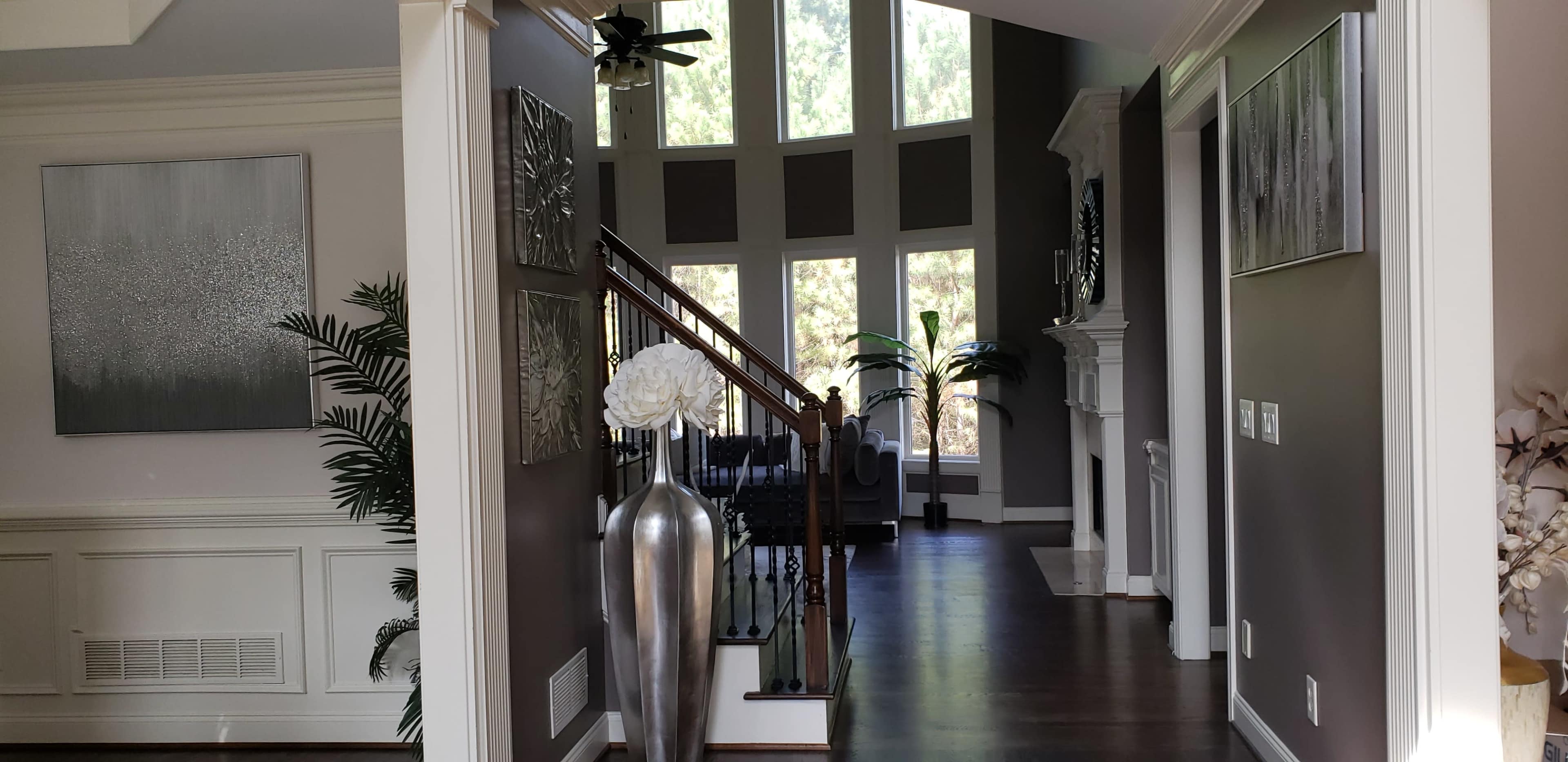 The image shows a spacious interior hallway leading to a sunlit living area, with tall windows, a staircase, and modern decor.