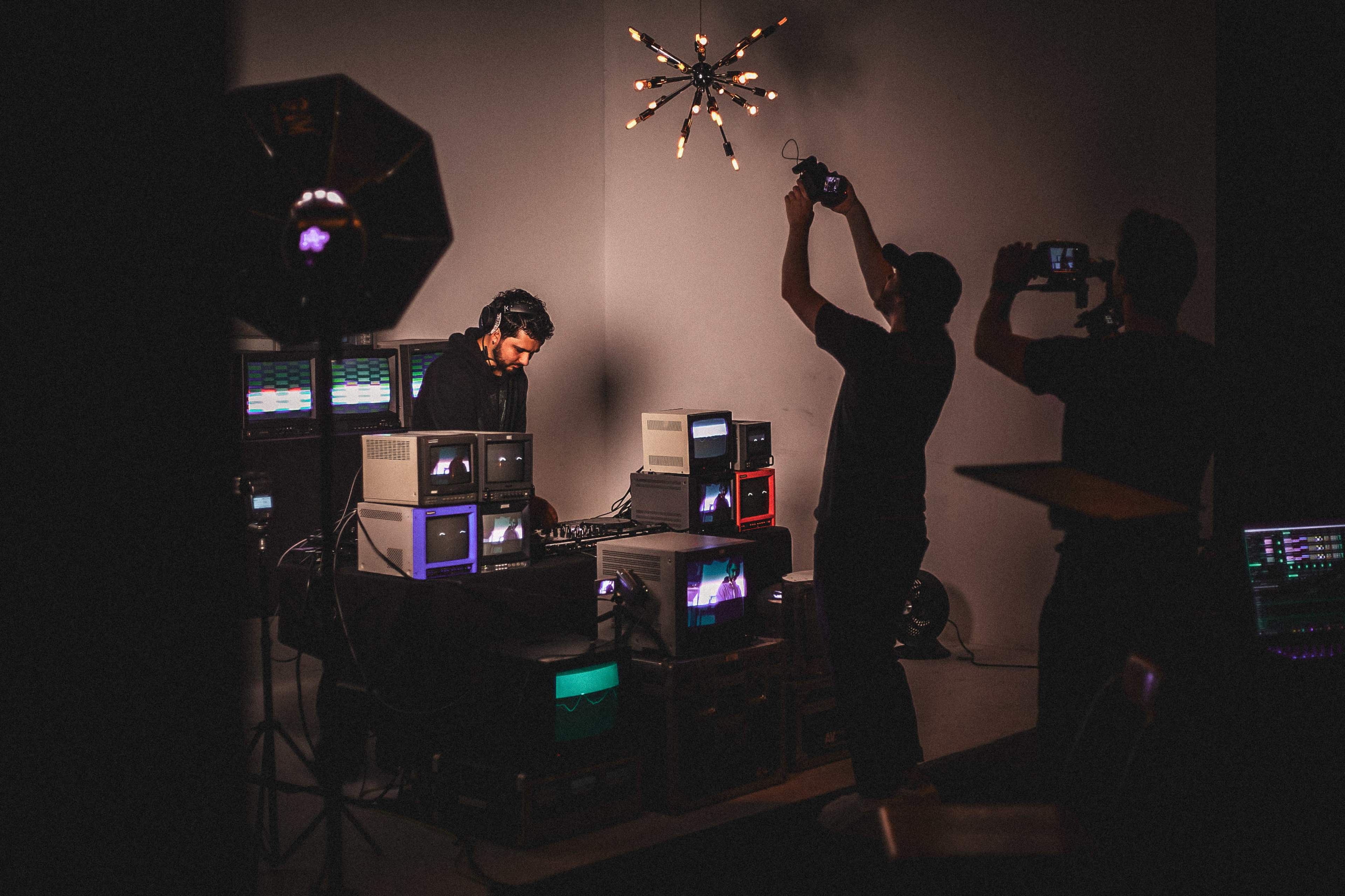 A musician is performing at a setup featuring retro computers and lighting, while two individuals record the scene using cameras.