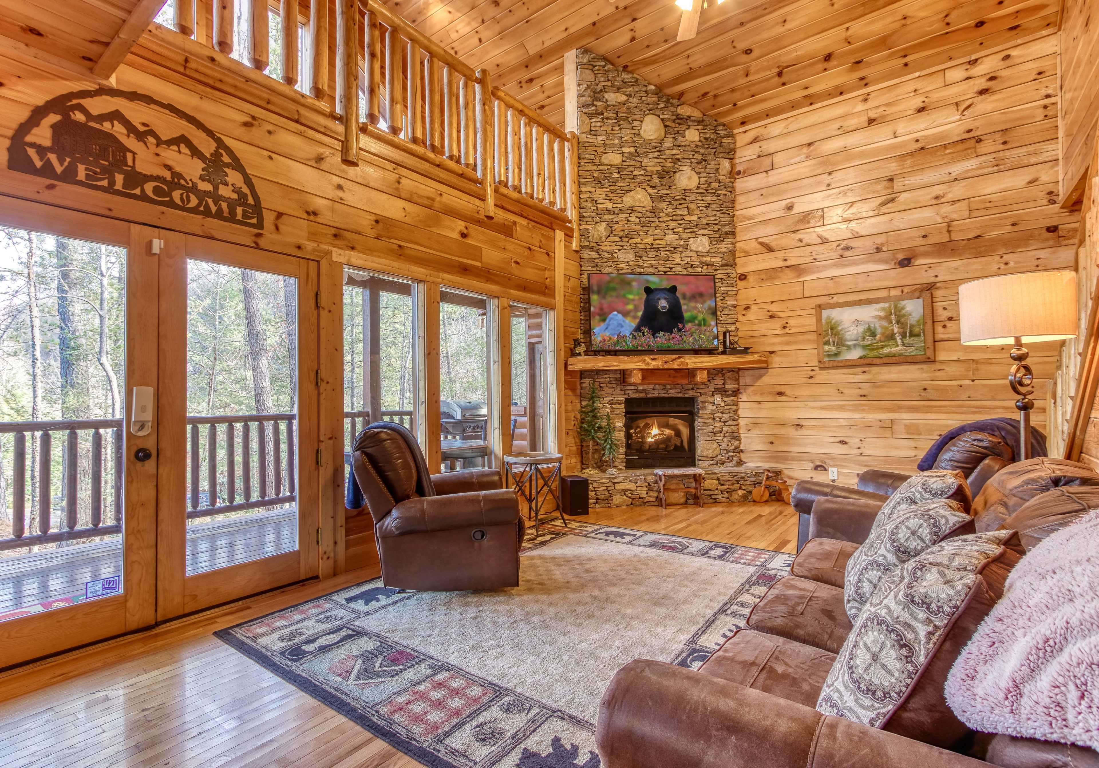The living room features wooden walls, a stone fireplace, a large window overlooking the forest, and comfortable seating.