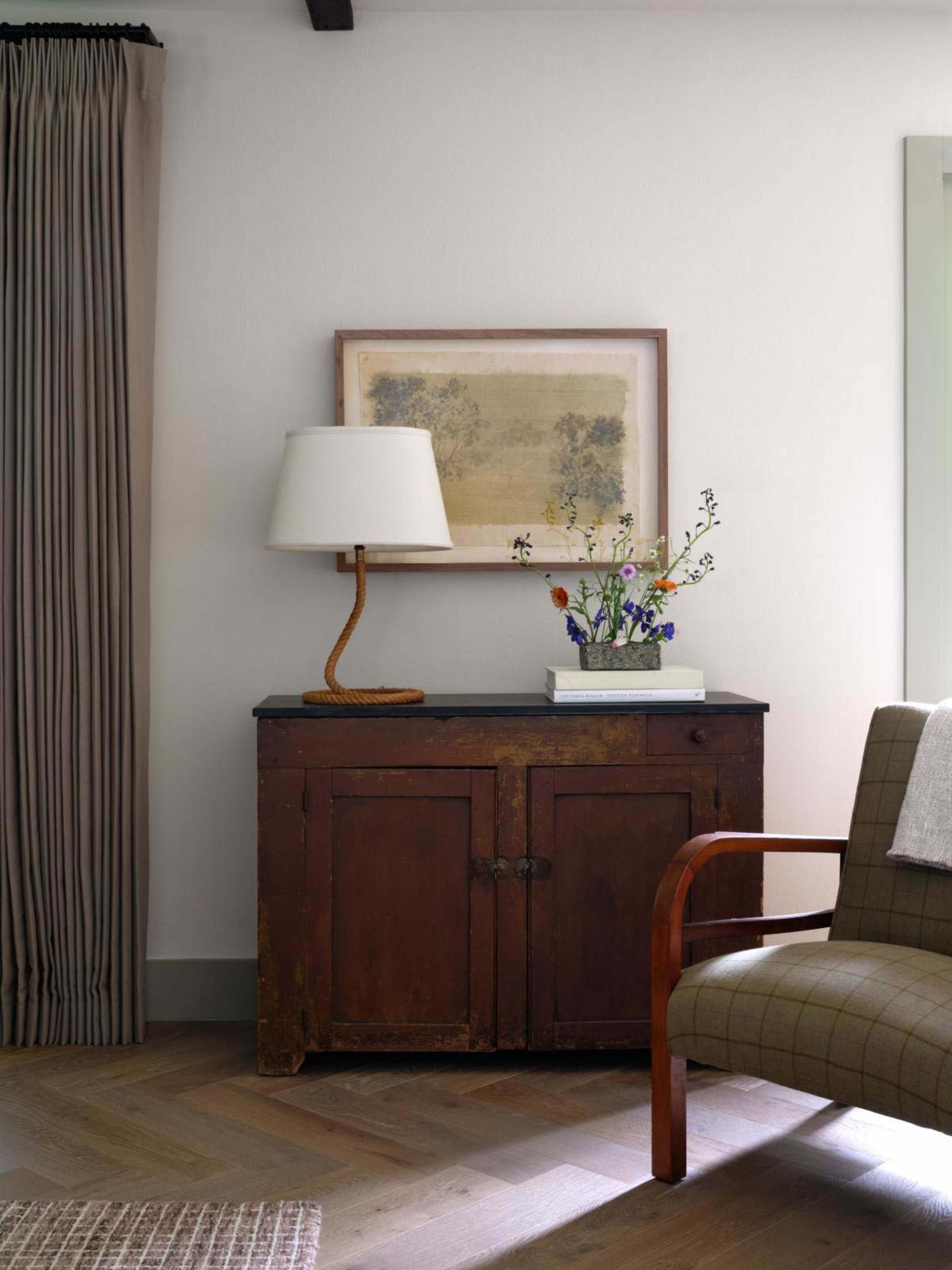 The image shows a wooden cabinet with a dark top and doors, a lamp, a vase of flowers, and a framed artwork, all positioned beside a chair and draped curtains.