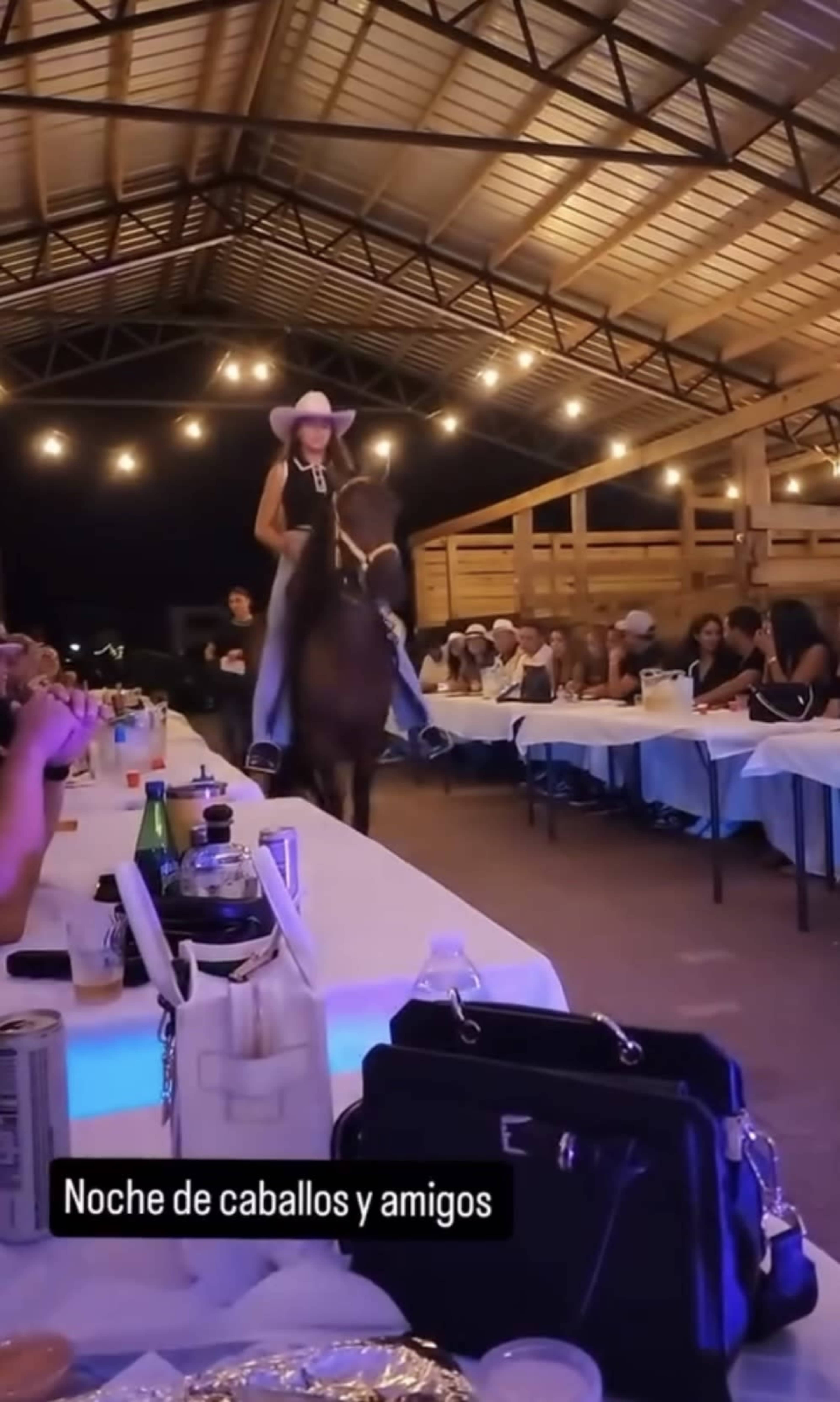 A woman in a cowboy hat rides a horse through a gathering of people seated at long tables under a lit barn structure.