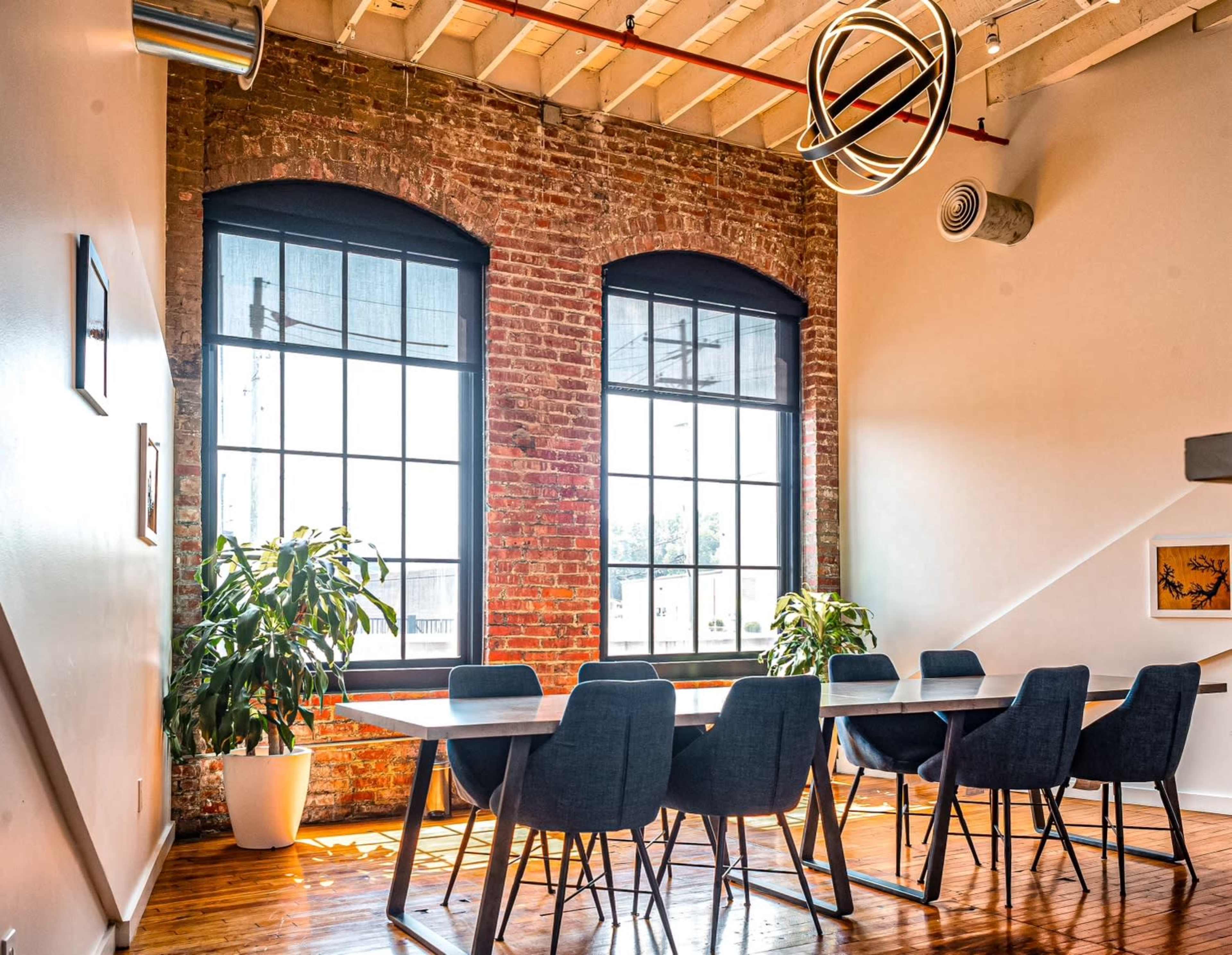 A brightly lit meeting room features a rectangular table surrounded by dark chairs, with large windows and exposed brick walls creating a modern aesthetic.