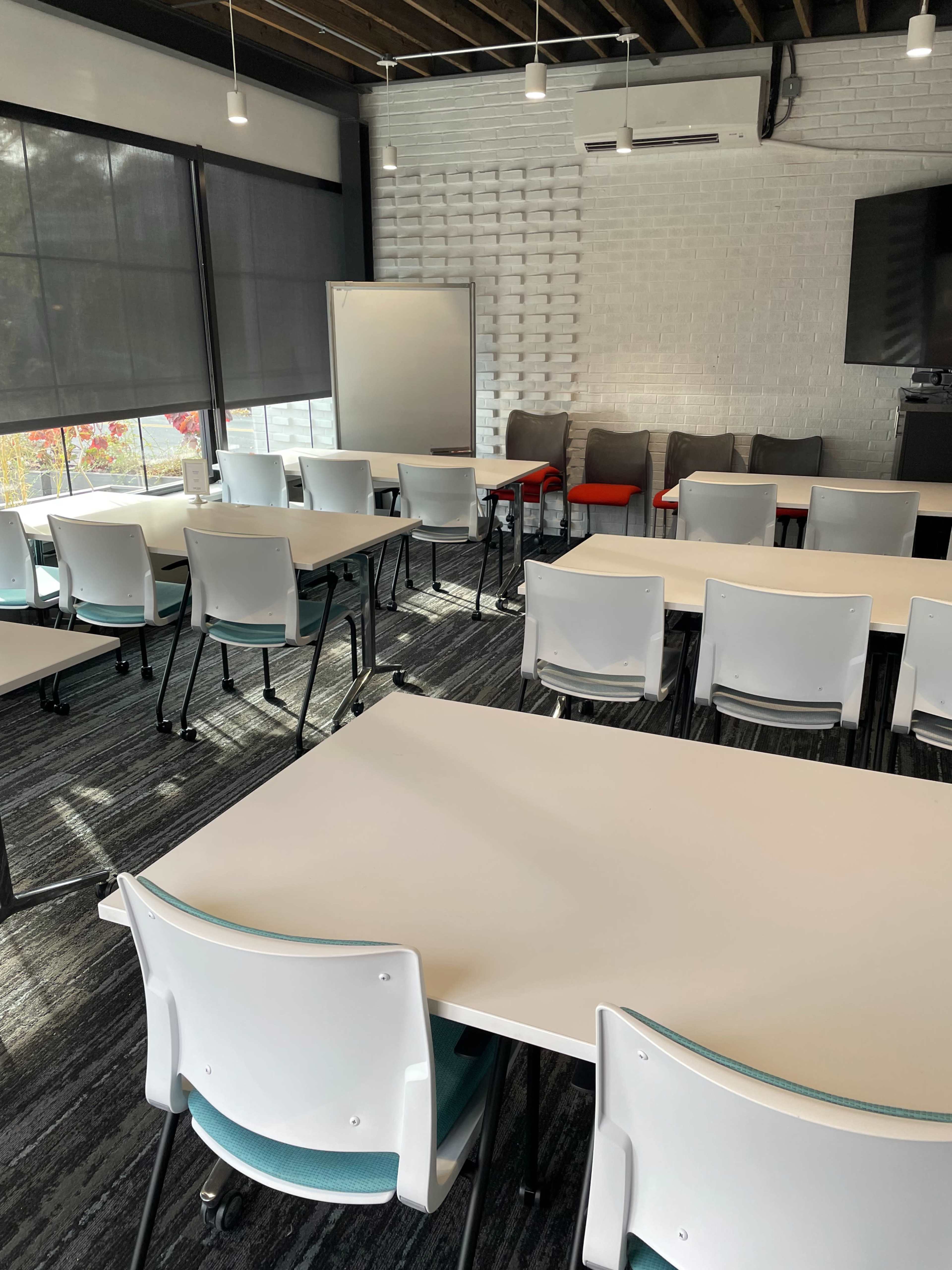 A modern conference room with several white tables arranged in a classroom style and chairs placed around them, complemented by a whiteboard and a seating area in the back.