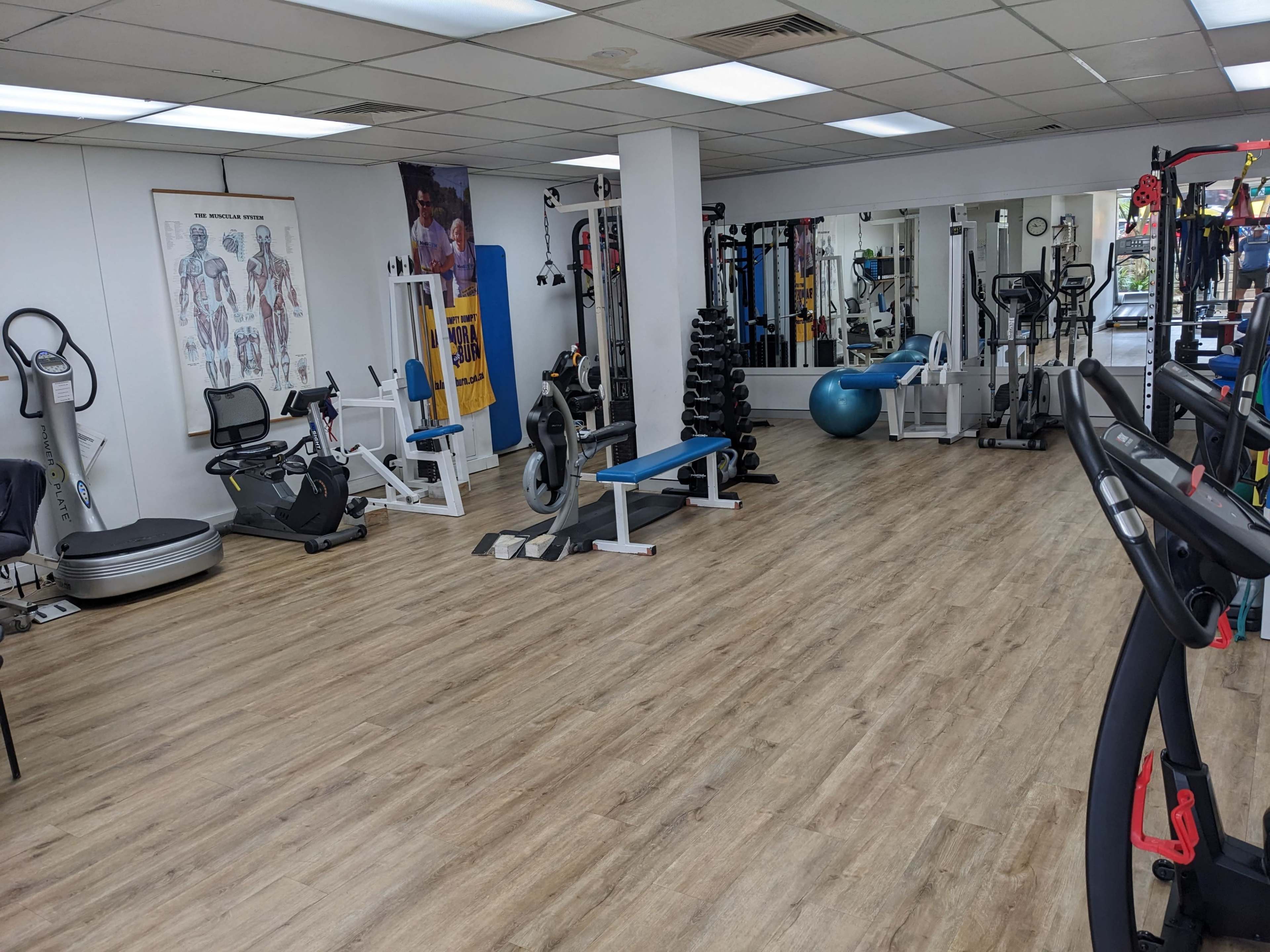 The image shows a fitness studio equipped with various exercise machines, weights, and a large mirror, featuring a wooden floor and bright lighting.