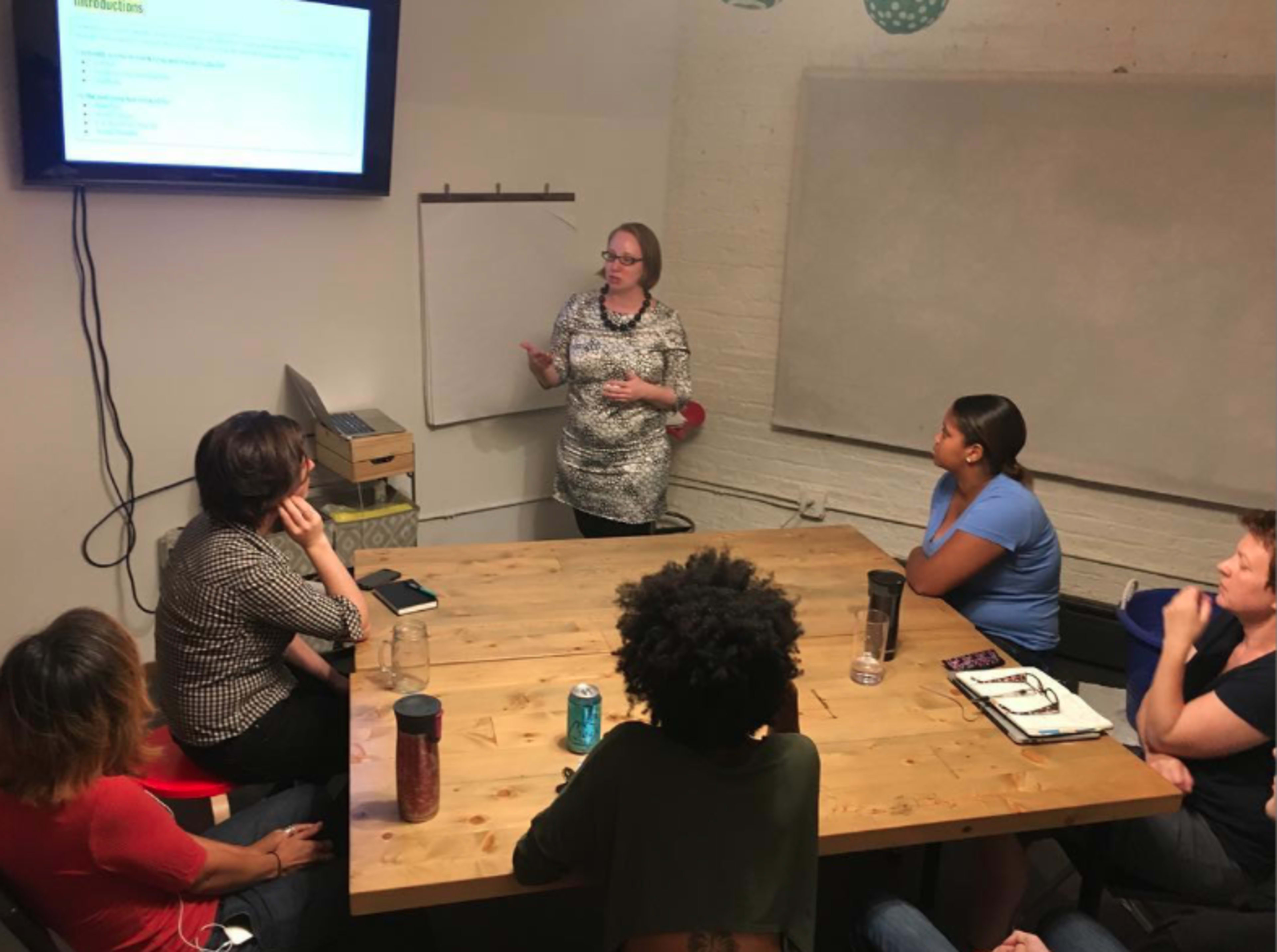 A woman presents information to a seated group of six people around a wooden table in a conference room.
