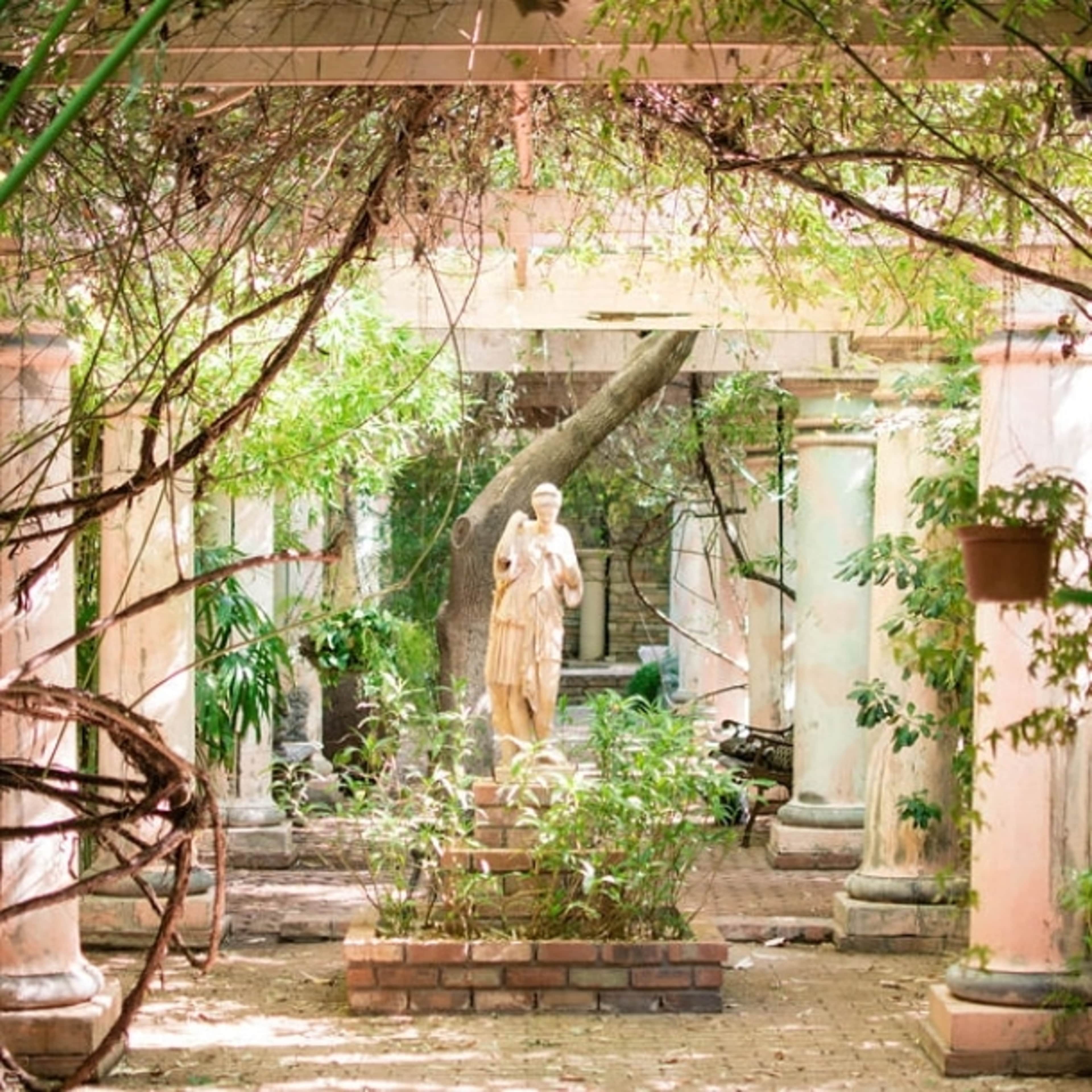 A statue stands amidst overgrown vegetation and weathered columns in a partially shaded garden.