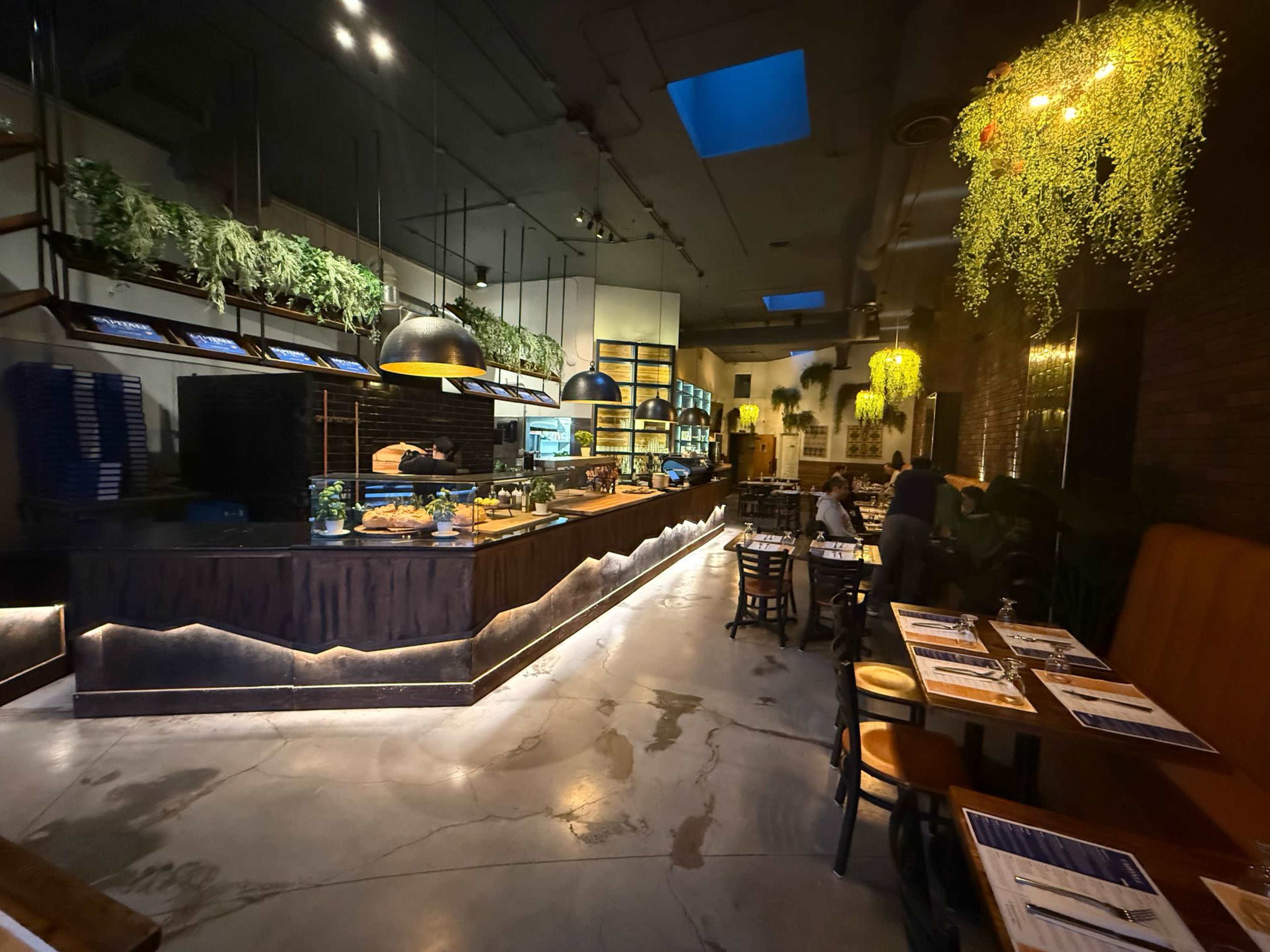 The image shows a modern restaurant interior with wooden furnishings, indoor plants, and ambient lighting, featuring a display counter and dining tables.