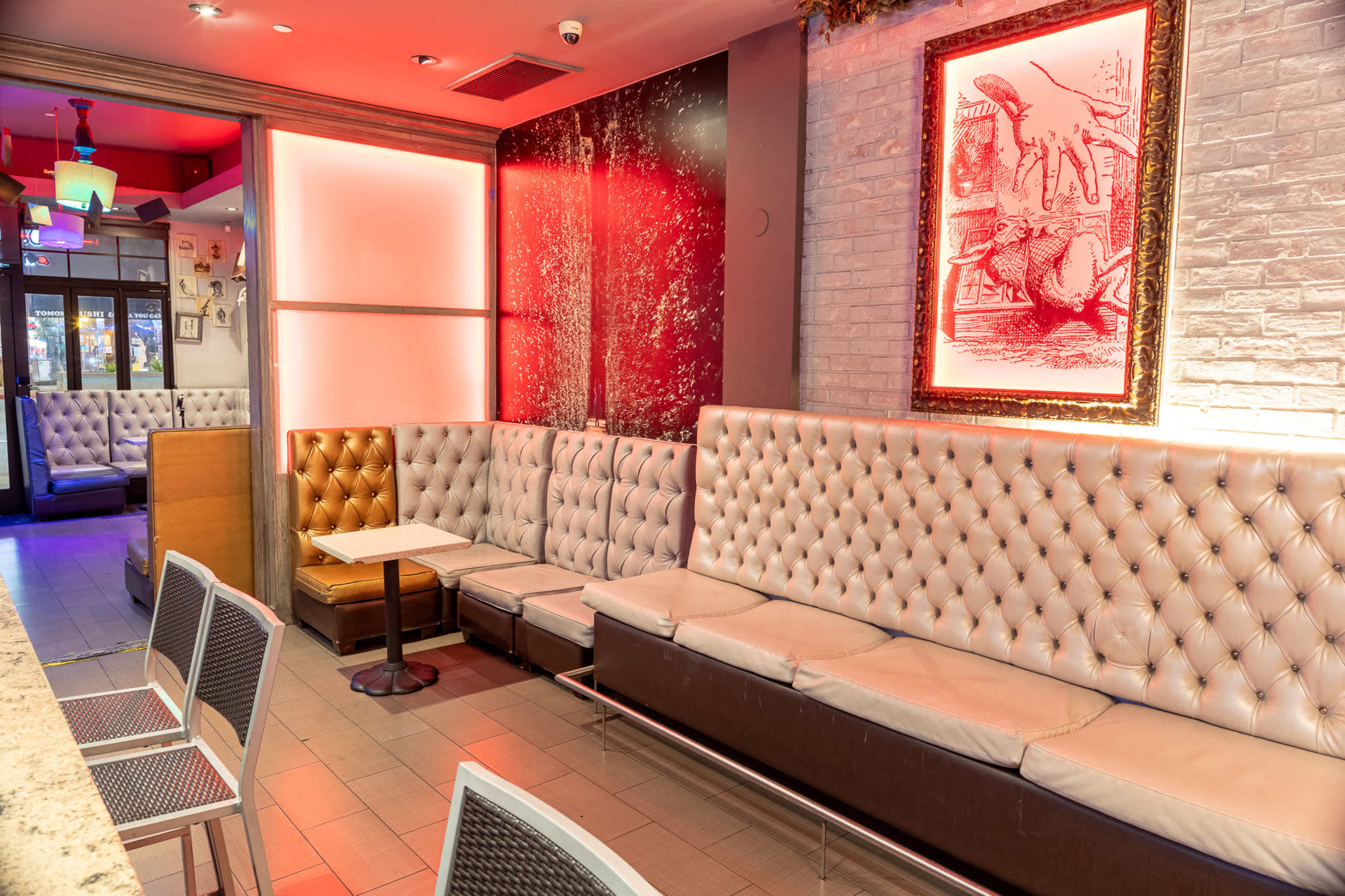 A modern seating area with tufted white benches, a small table, and a framed artwork on a brick wall, illuminated by red lighting.