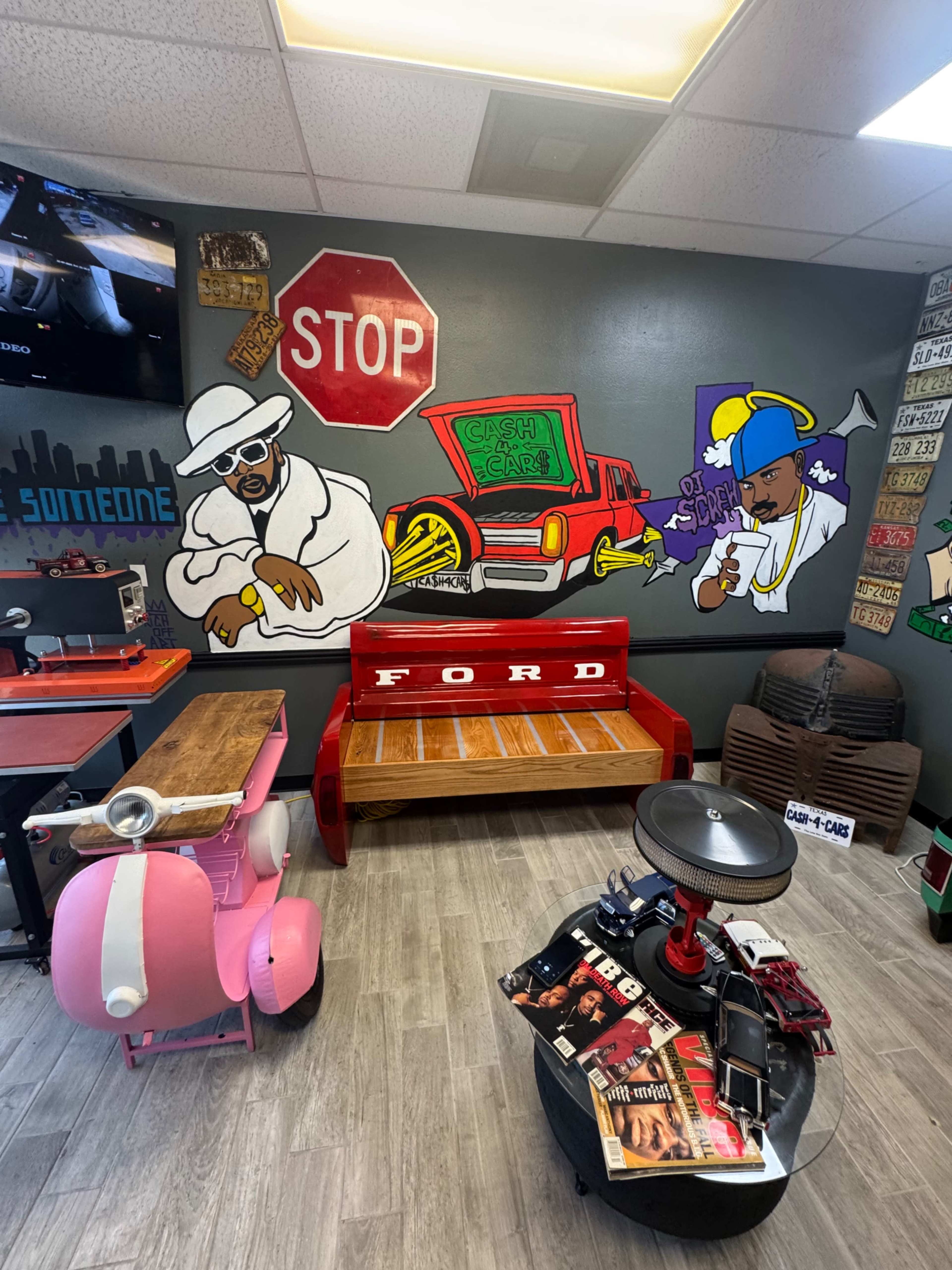 The image shows a colorful wall mural featuring two cartoon figures alongside a vintage Ford bed, with a pink scooter and various items scattered around the room.
