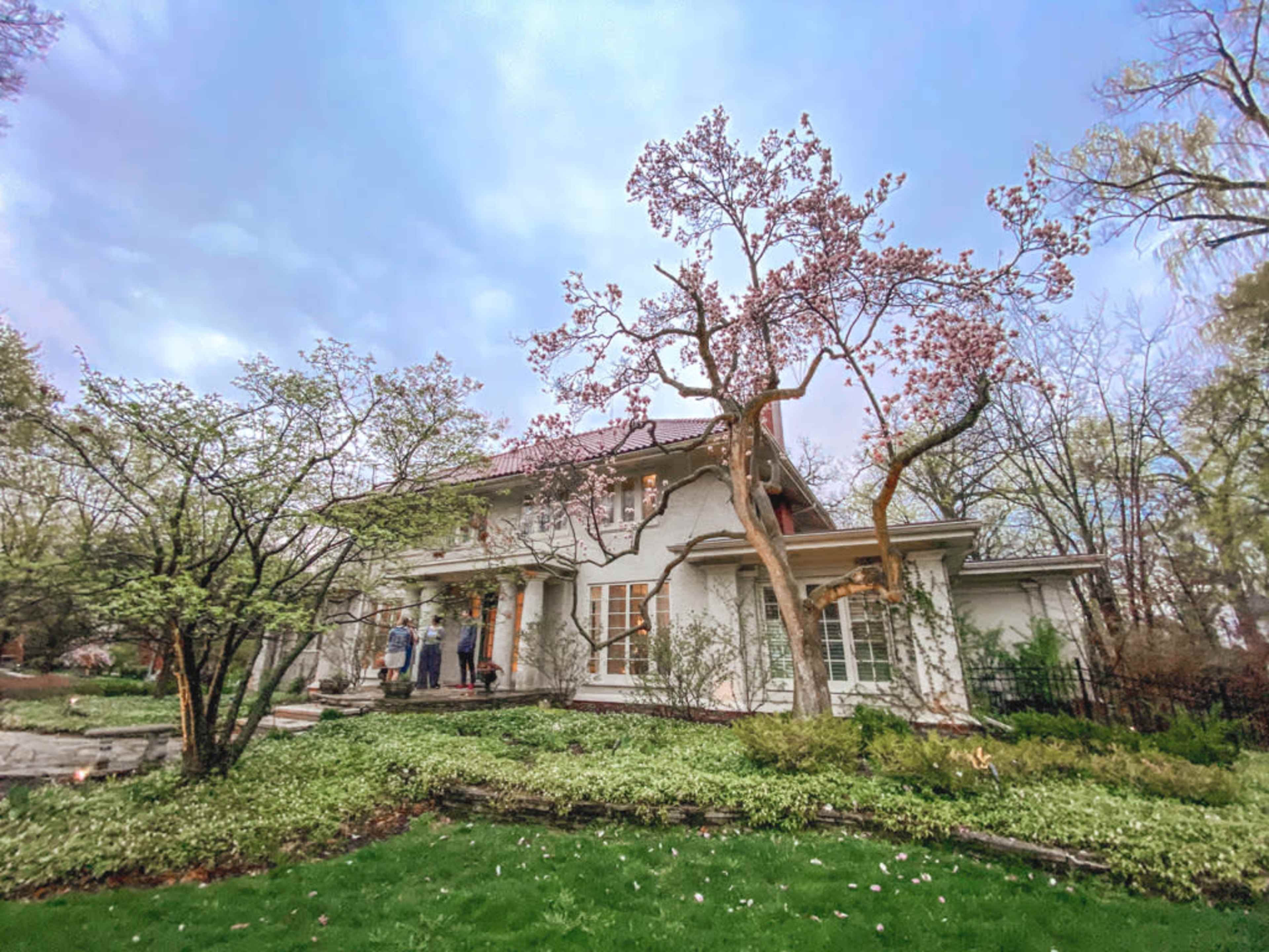 A white house with a covered porch is surrounded by greenery and a flowering tree, set against a cloudy sky.