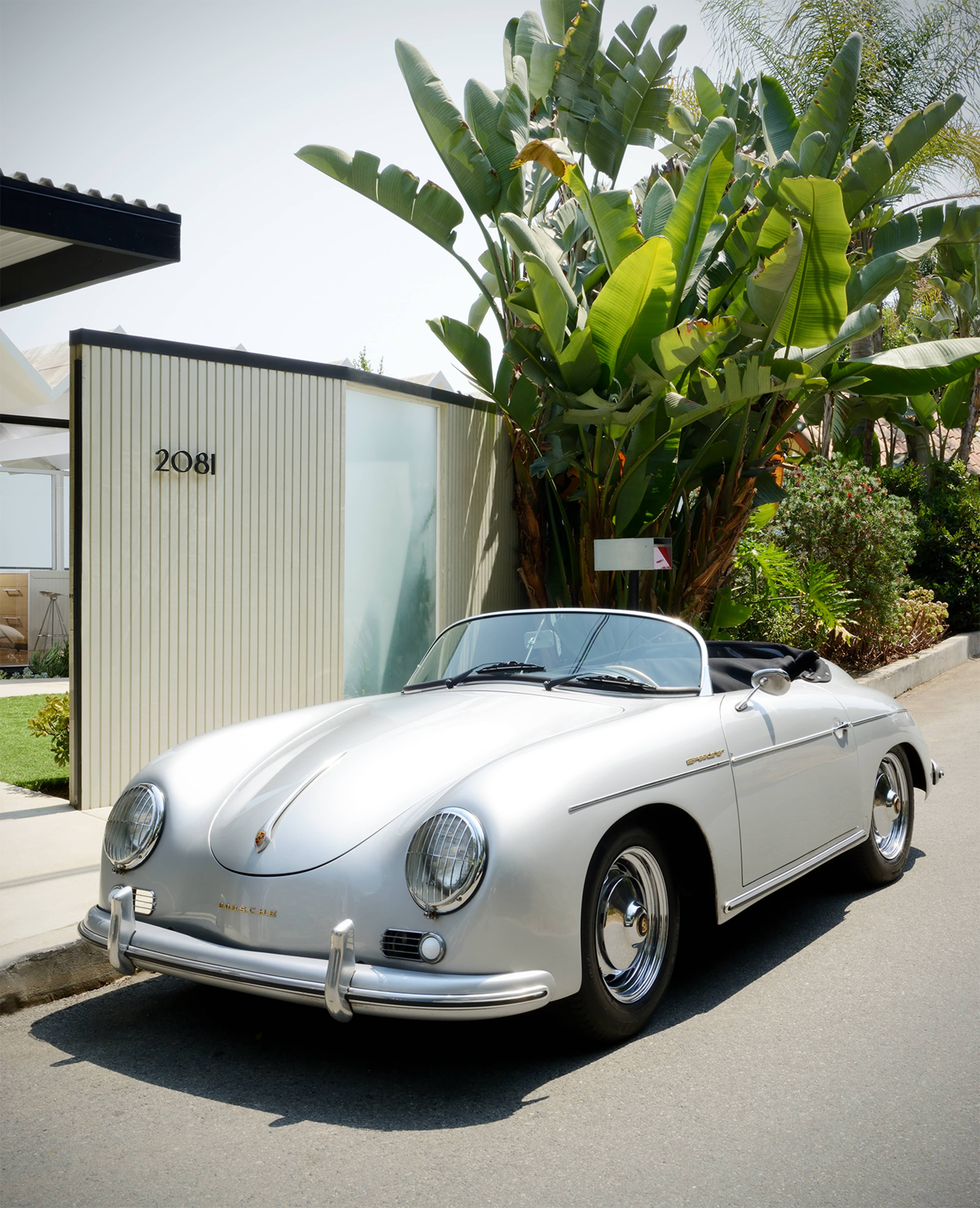 A silver convertible sports car is parked beside a modern building with lush greenery in a sunny residential area.