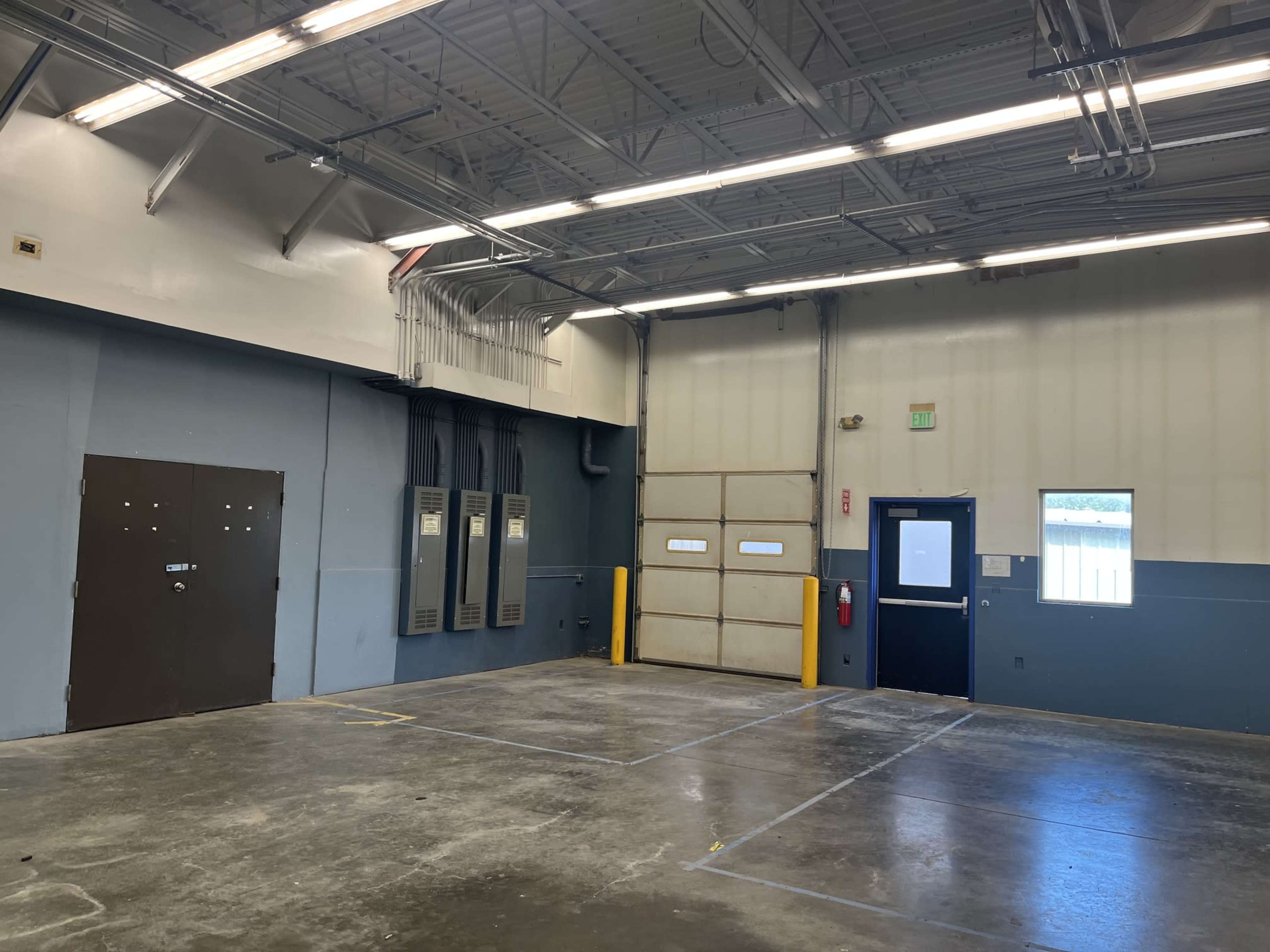 The image shows an industrial space with a concrete floor, blue and white walls, a large roll-up door, and electrical panels on one side.