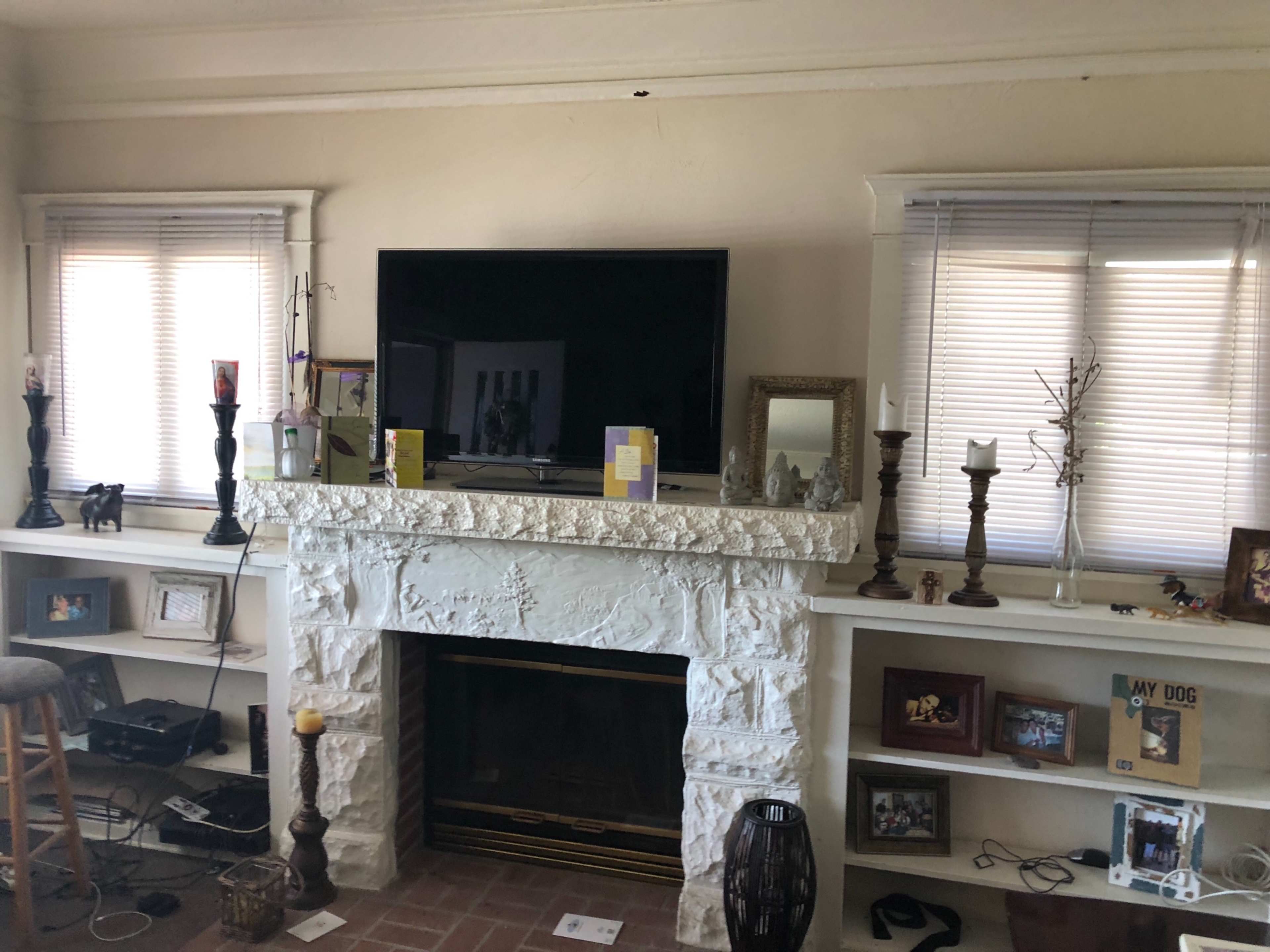 A living room with a stone fireplace underneath a mounted television, flanked by two windows with blinds, and shelves displaying various decorative items and family photos.