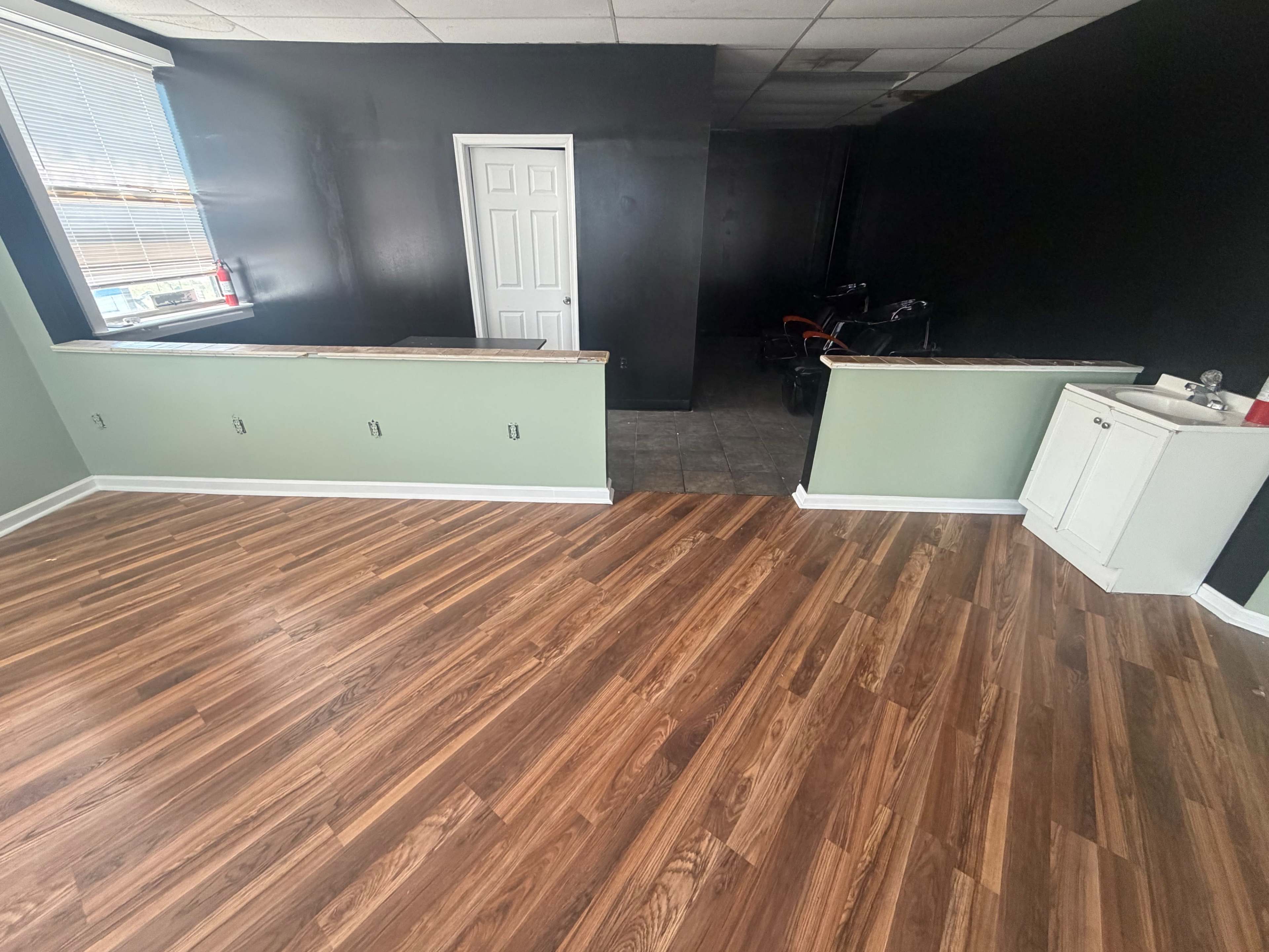 The image shows a room with light green walls, a wooden floor, and a kitchenette area with a white cabinet in the corner, while the adjacent area features black walls.