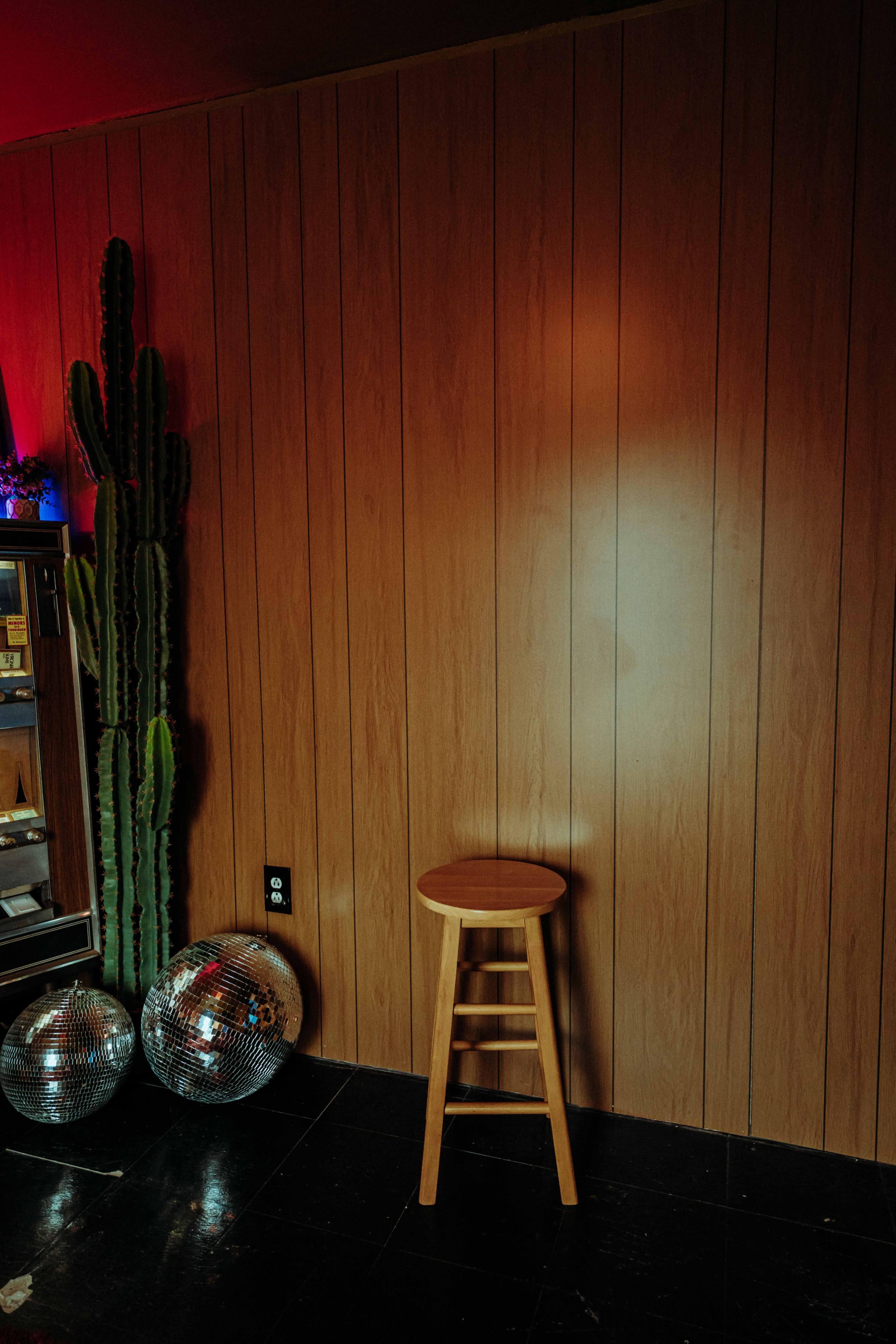 A wooden stool stands against a wooden wall next to two disco balls and a tall cactus.