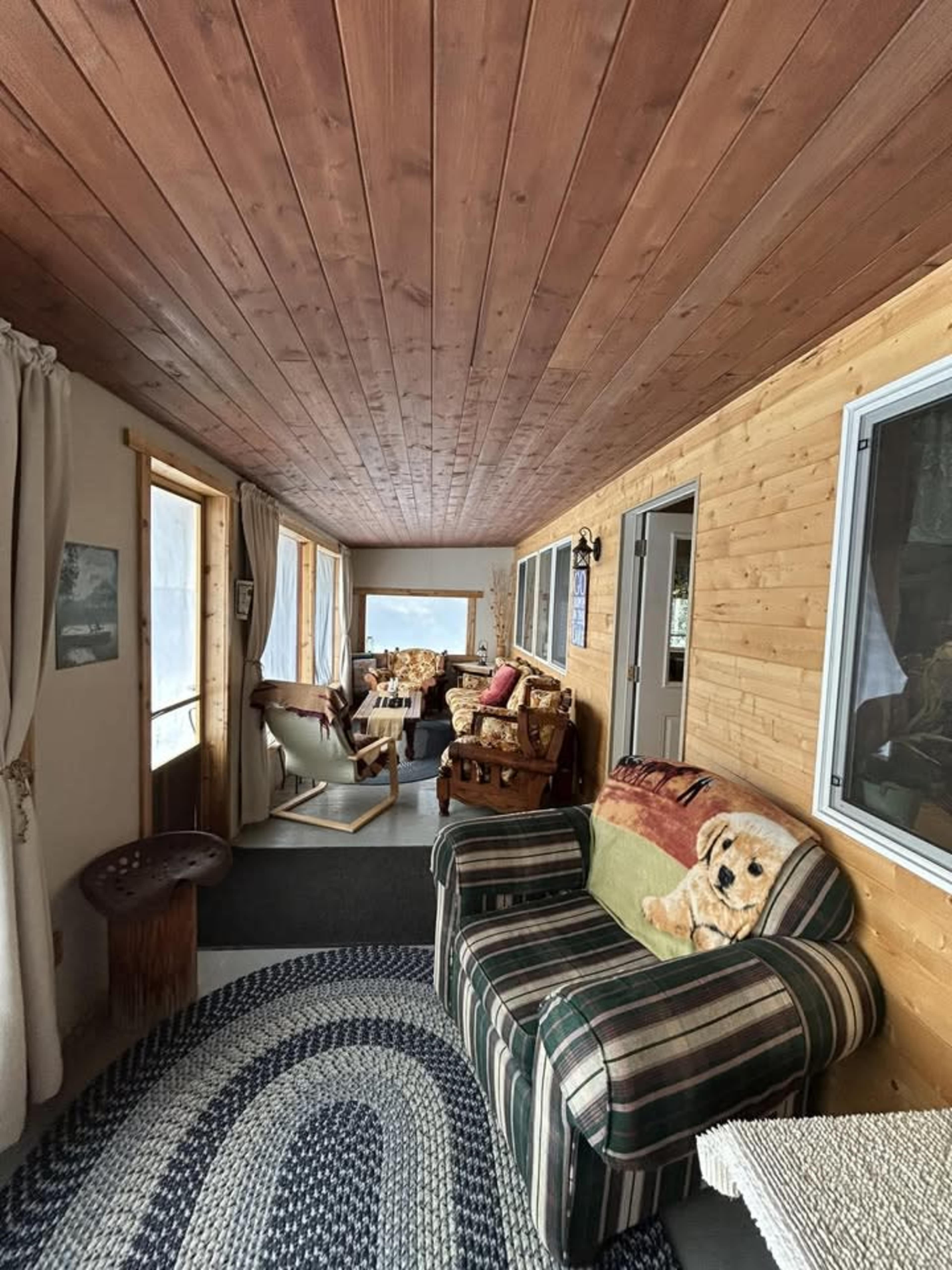 The image shows a cozy enclosed porch featuring wooden walls, a striped couch, and various seating arrangements with large windows allowing natural light to enter.