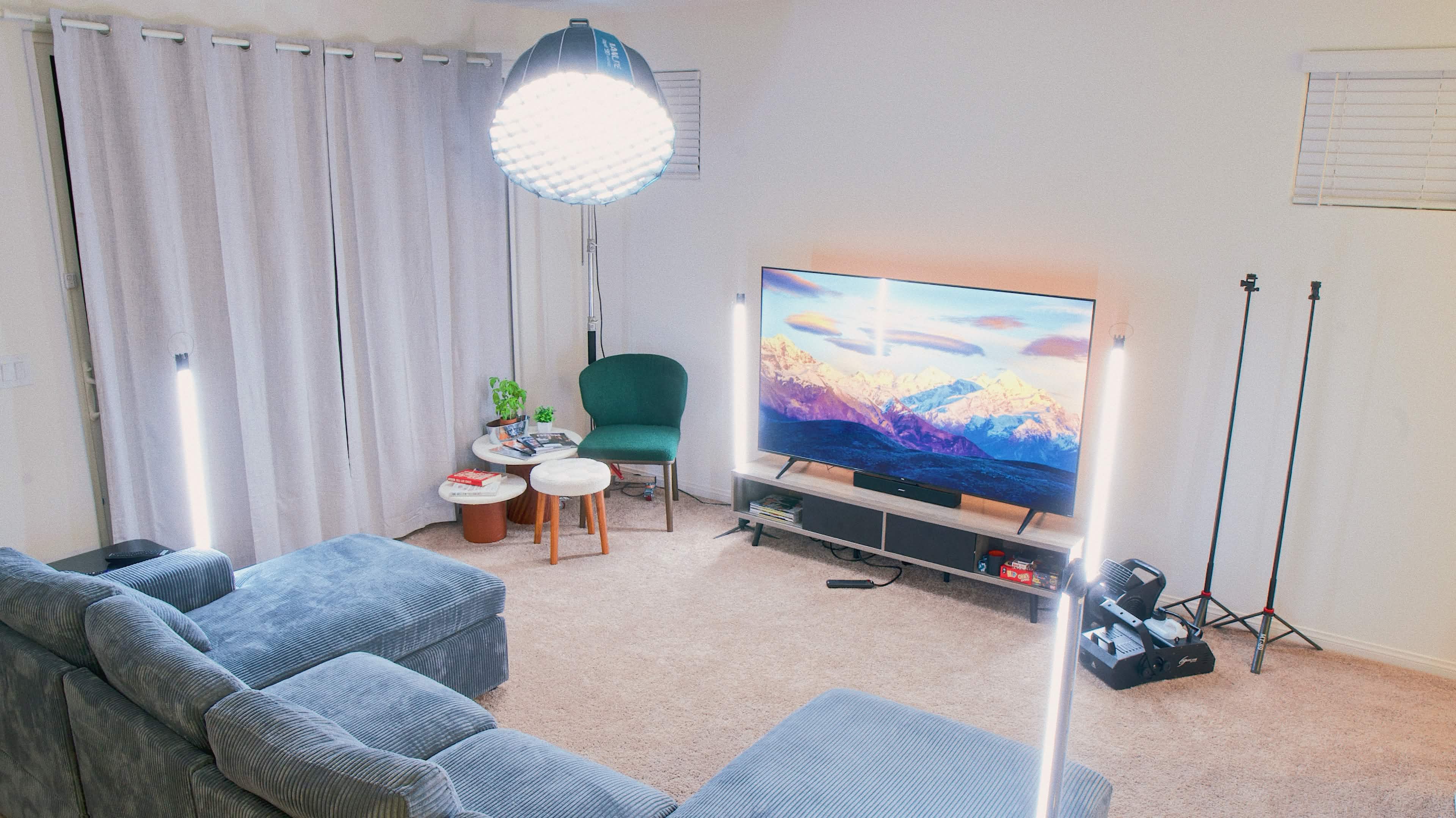 The living room features a large television displaying a mountain landscape, surrounded by a sectional sofa, lighting equipment, and a small table with plants.