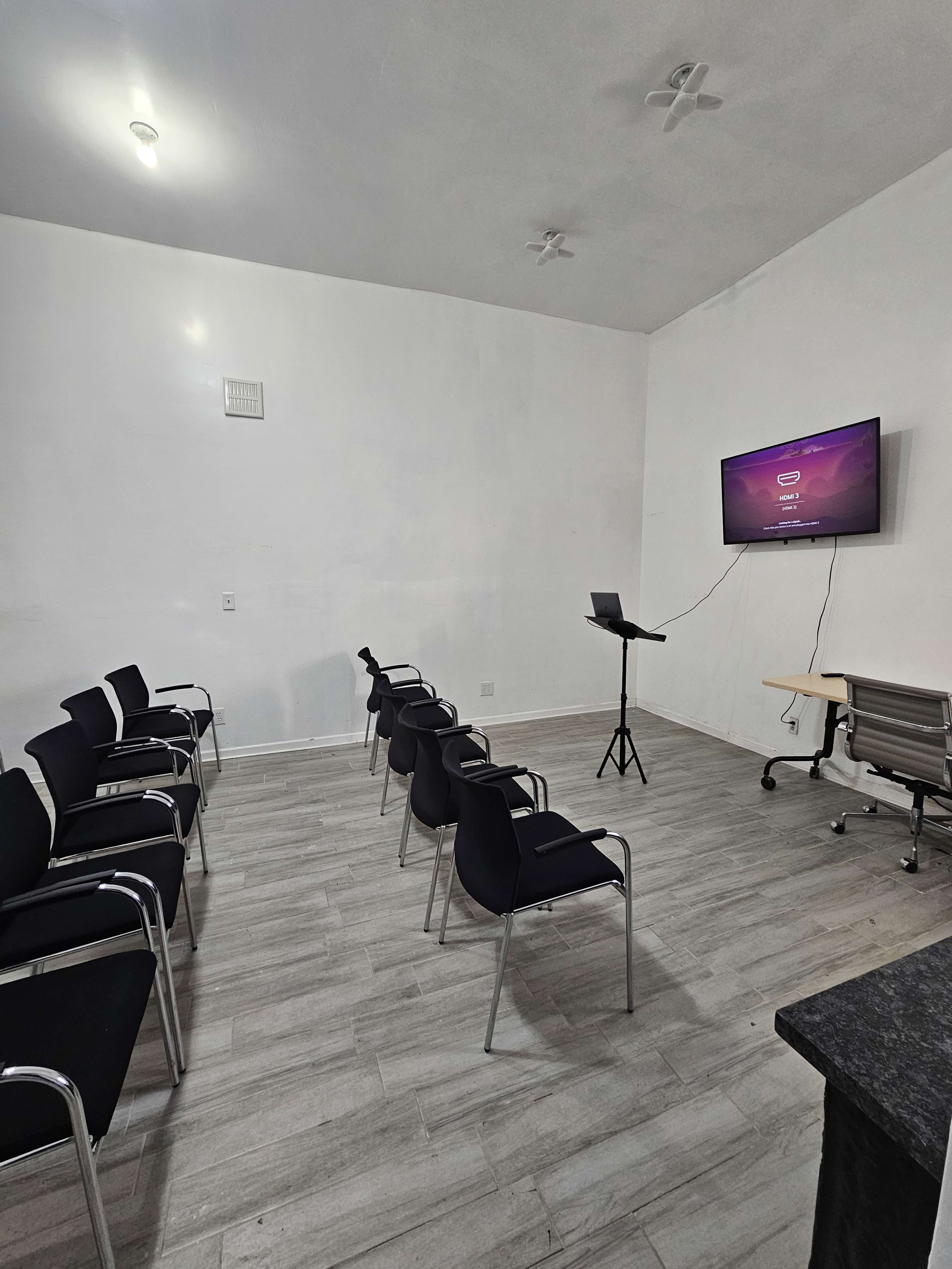 The image shows a small meeting room with several black chairs arranged in rows facing a screen mounted on the wall.
