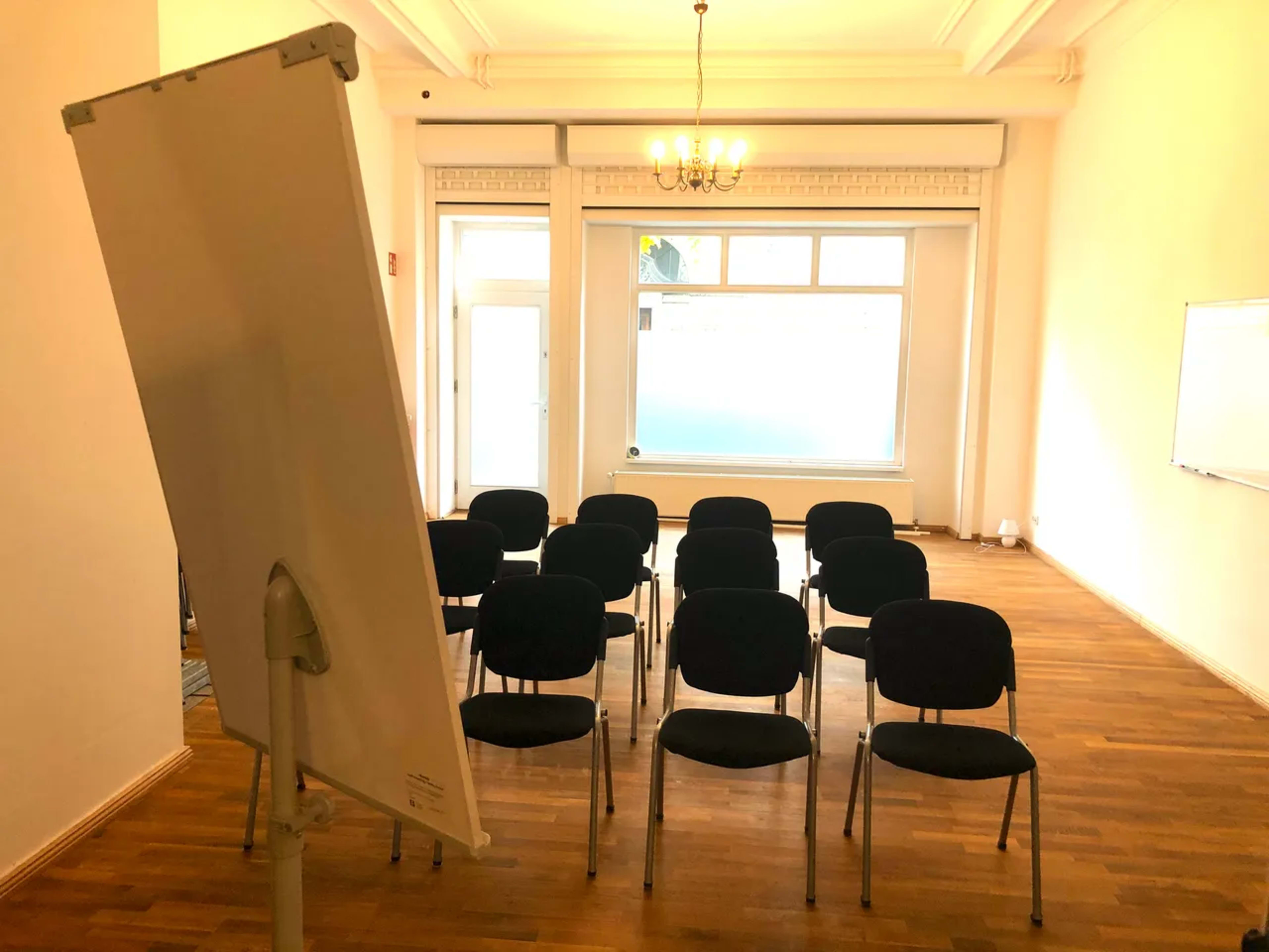 A spacious room with wooden flooring contains several chairs arranged in rows facing a flip chart and a large window.