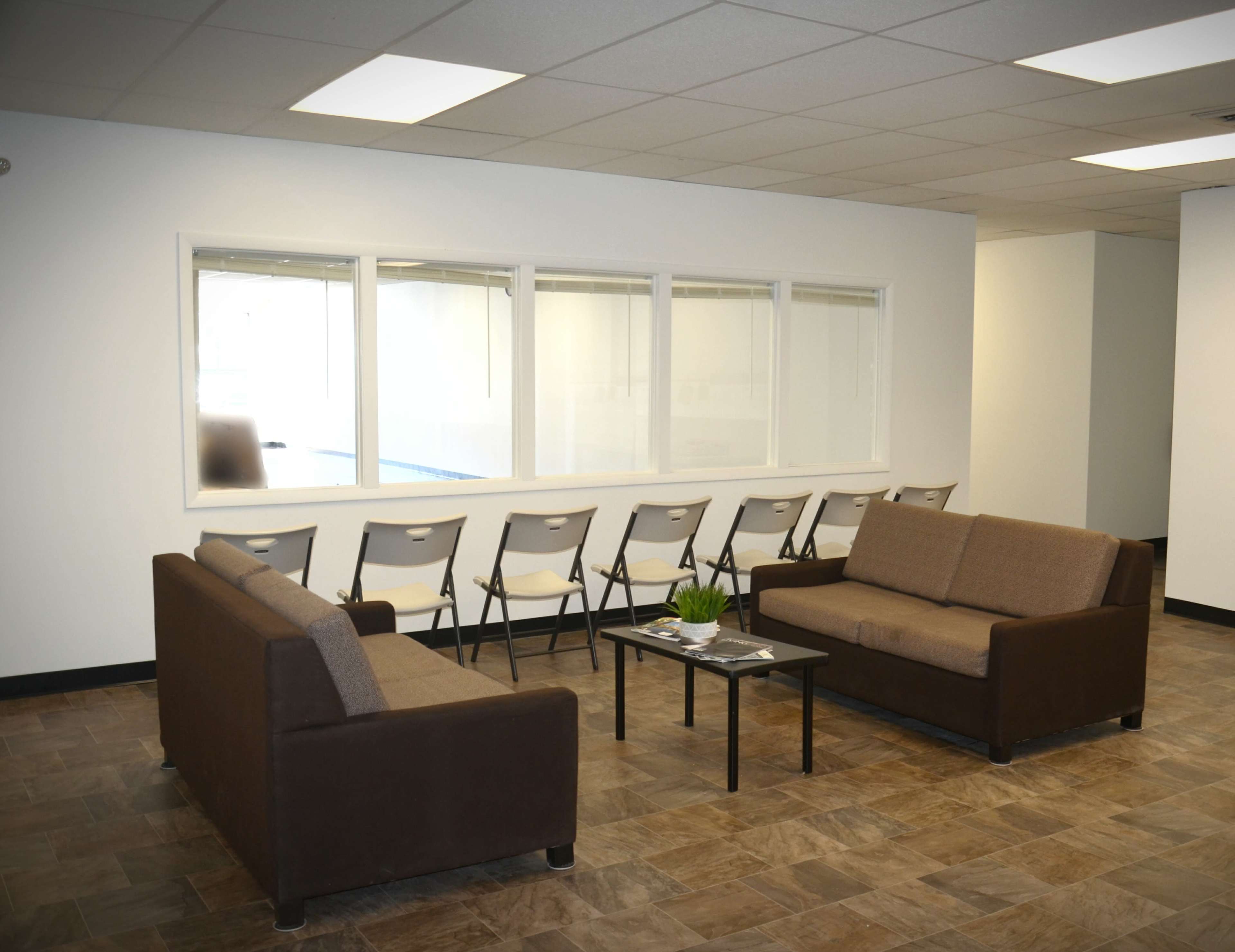 A waiting area with two brown sofas, a small table, and a row of folding chairs positioned against a wall with large windows.