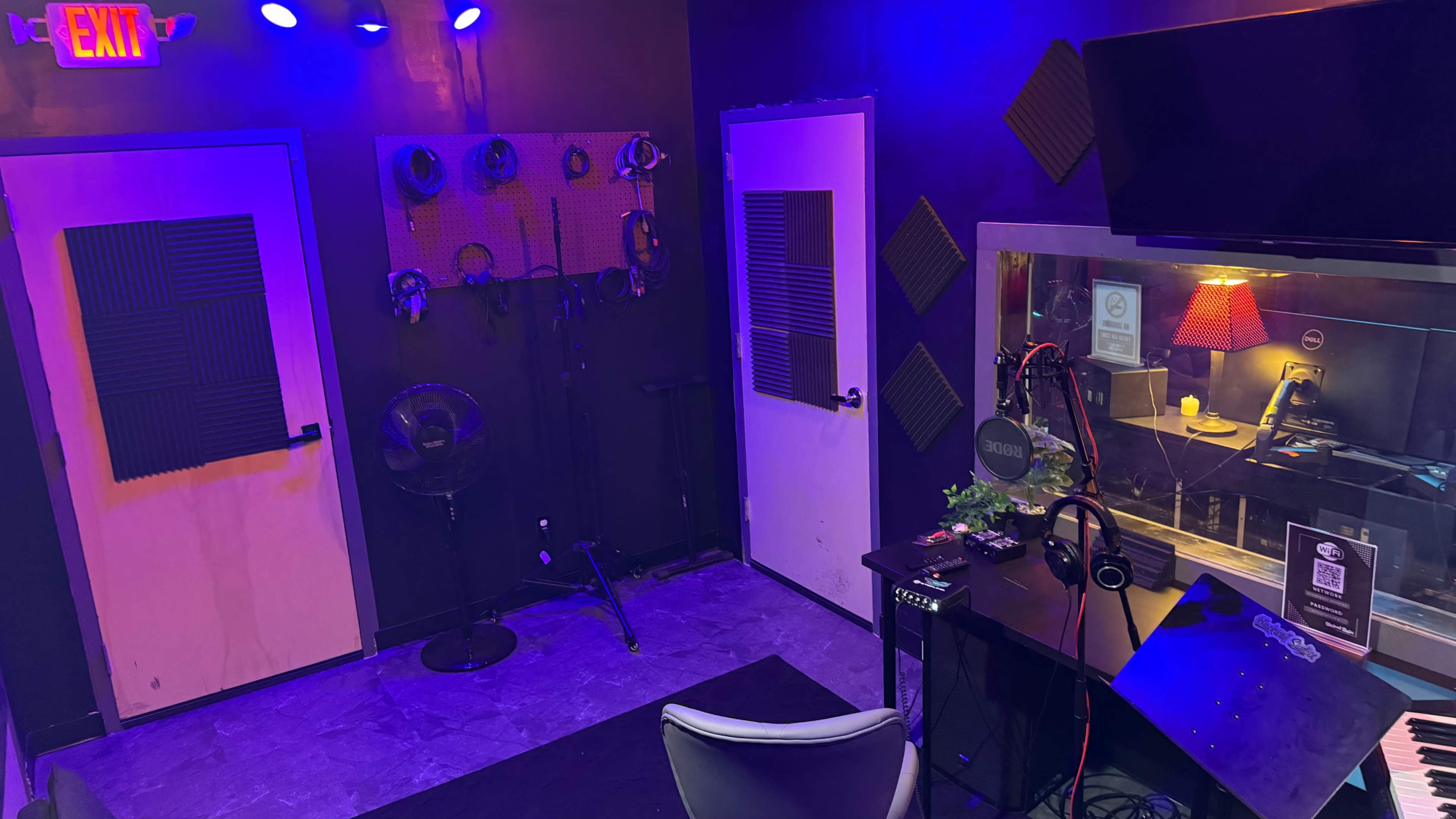The image shows a small recording studio with two white doors, a microphone setup, headphones, and soundproofing materials on the walls, all illuminated by blue and purple lighting.