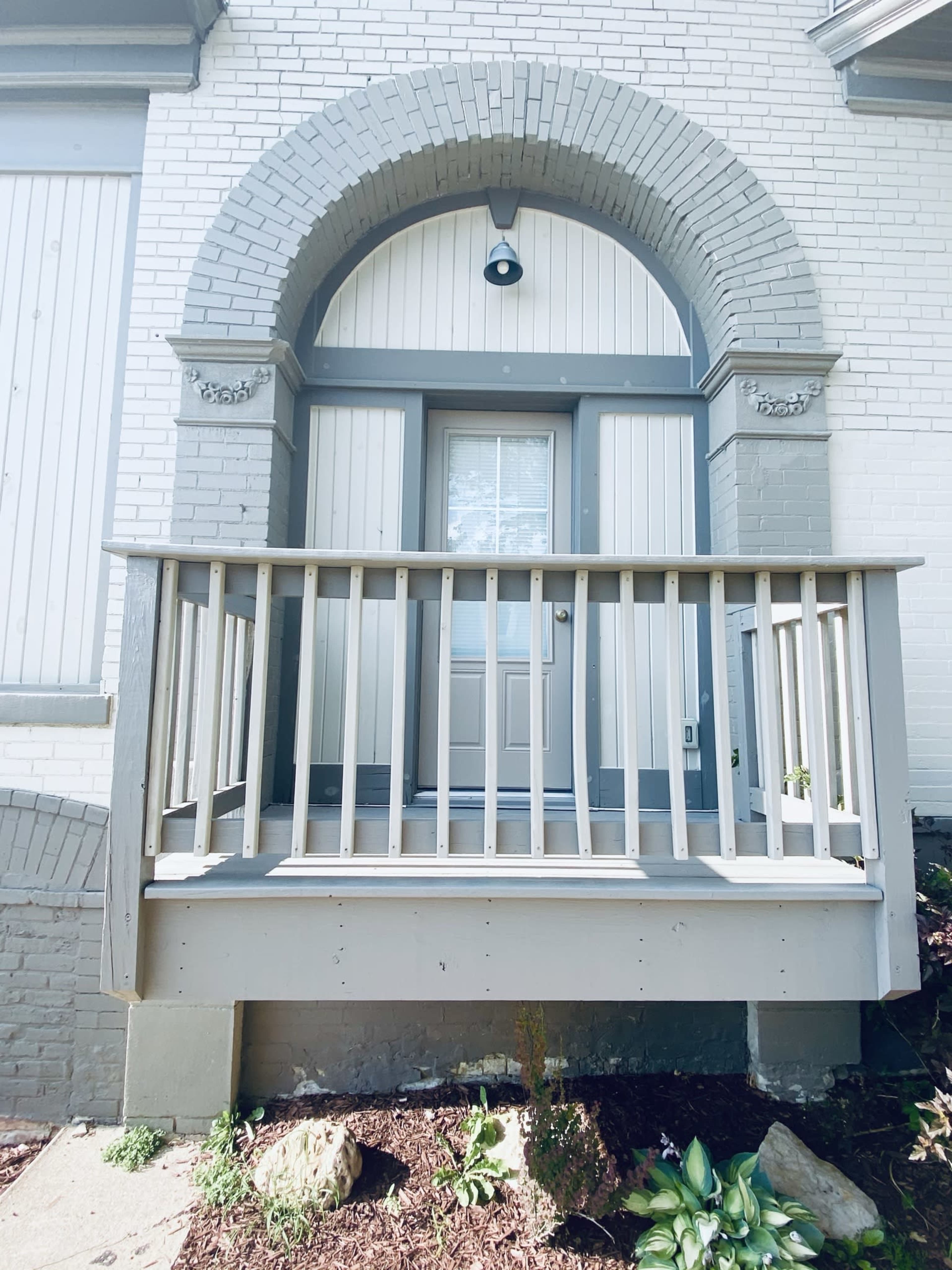 A small front porch with a wooden railing leading up to a door framed by an arched structure on a brick building.