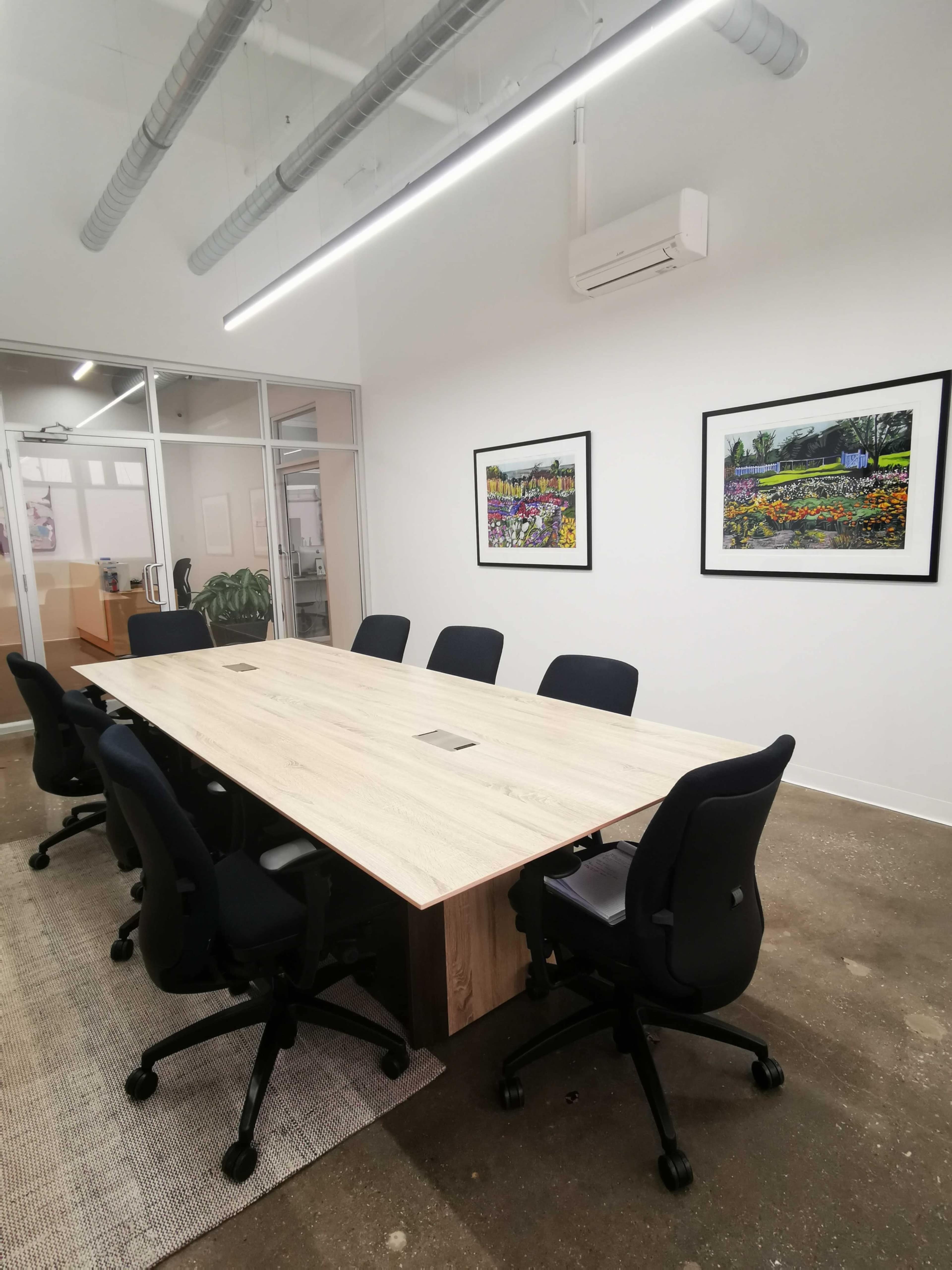 A long wooden conference table is surrounded by black office chairs in a well-lit meeting room with framed artwork on the walls.