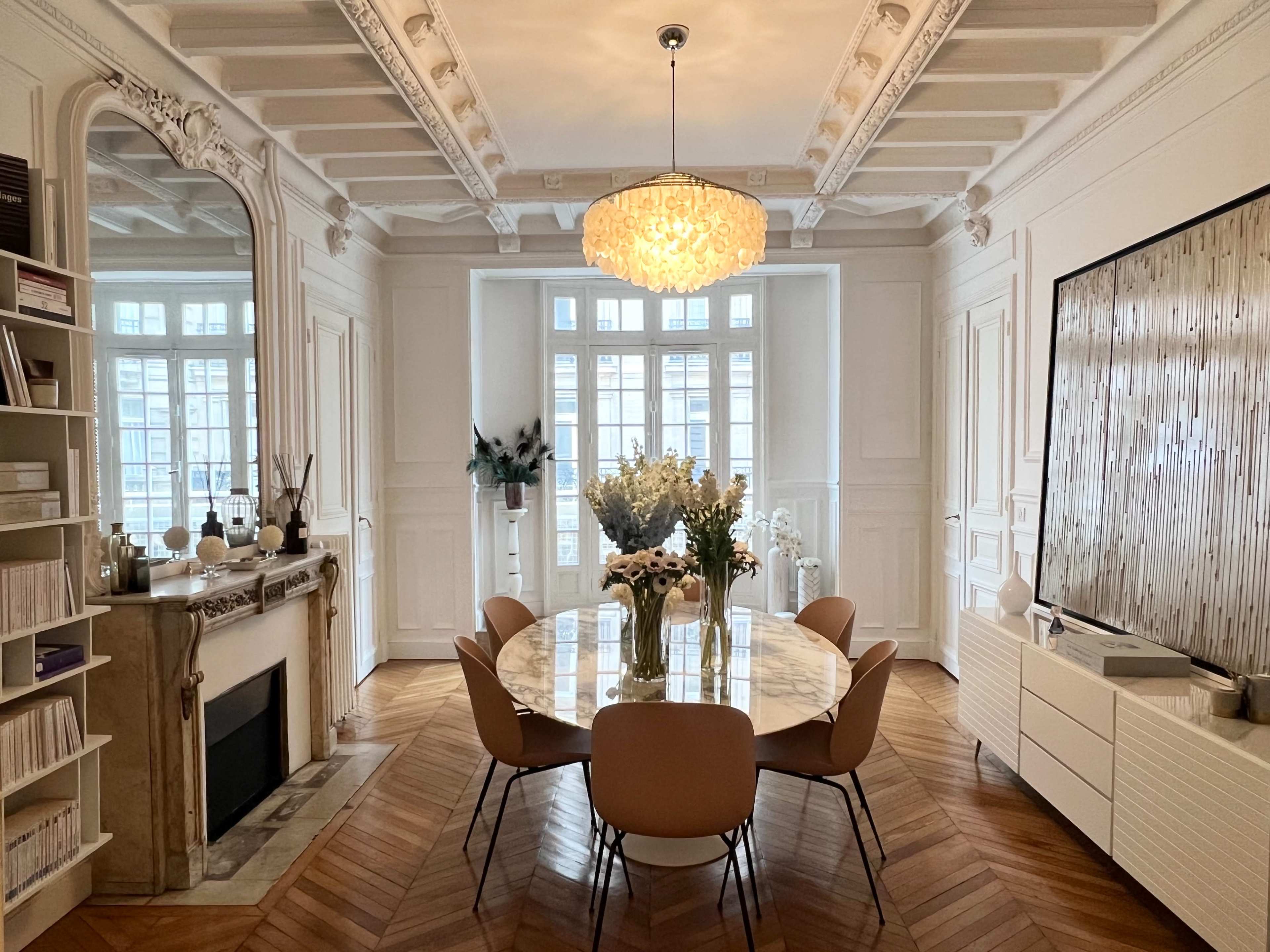 A modern dining room features a round marble table surrounded by beige chairs, with a large chandelier overhead and decorative elements on the walls.