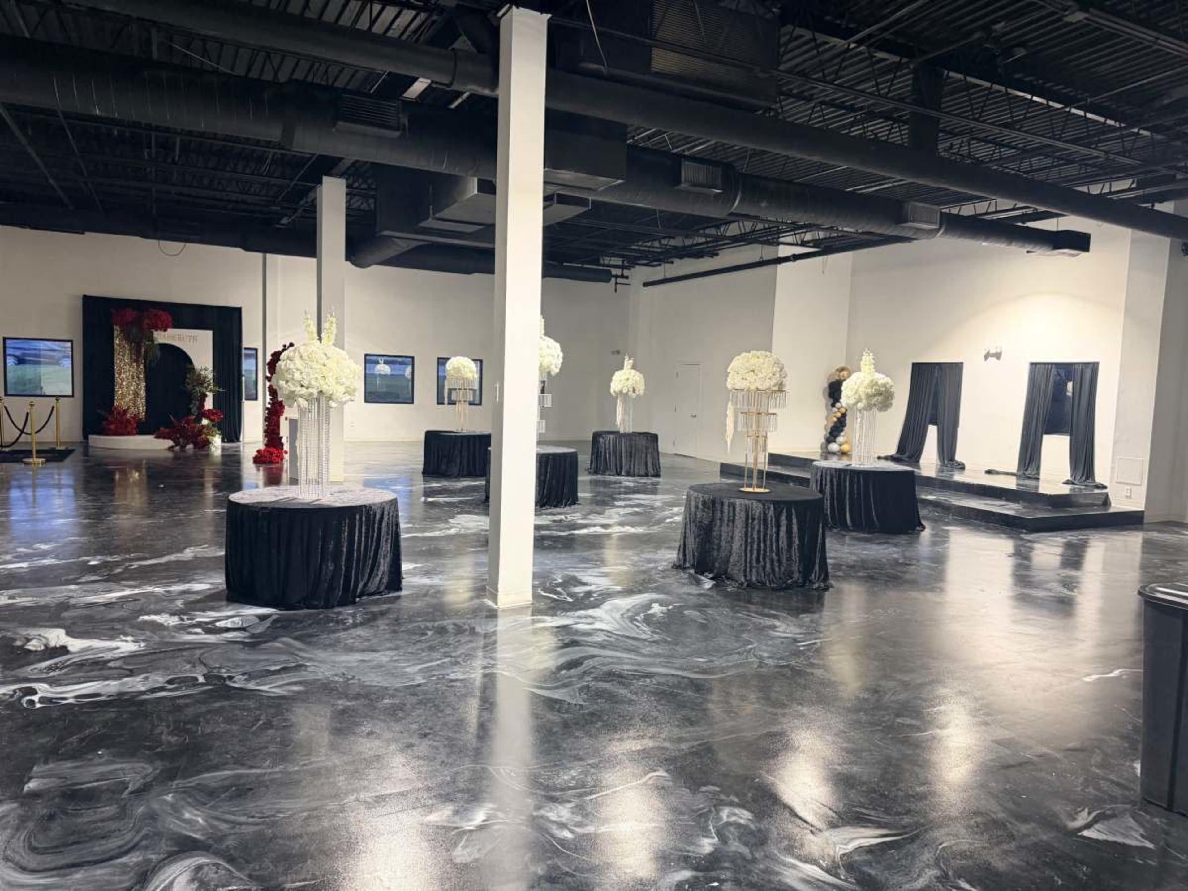 The image shows a spacious indoor venue featuring several round tables adorned with floral centerpieces, a polished floor with a marble-like pattern, and decorative elements along the walls.