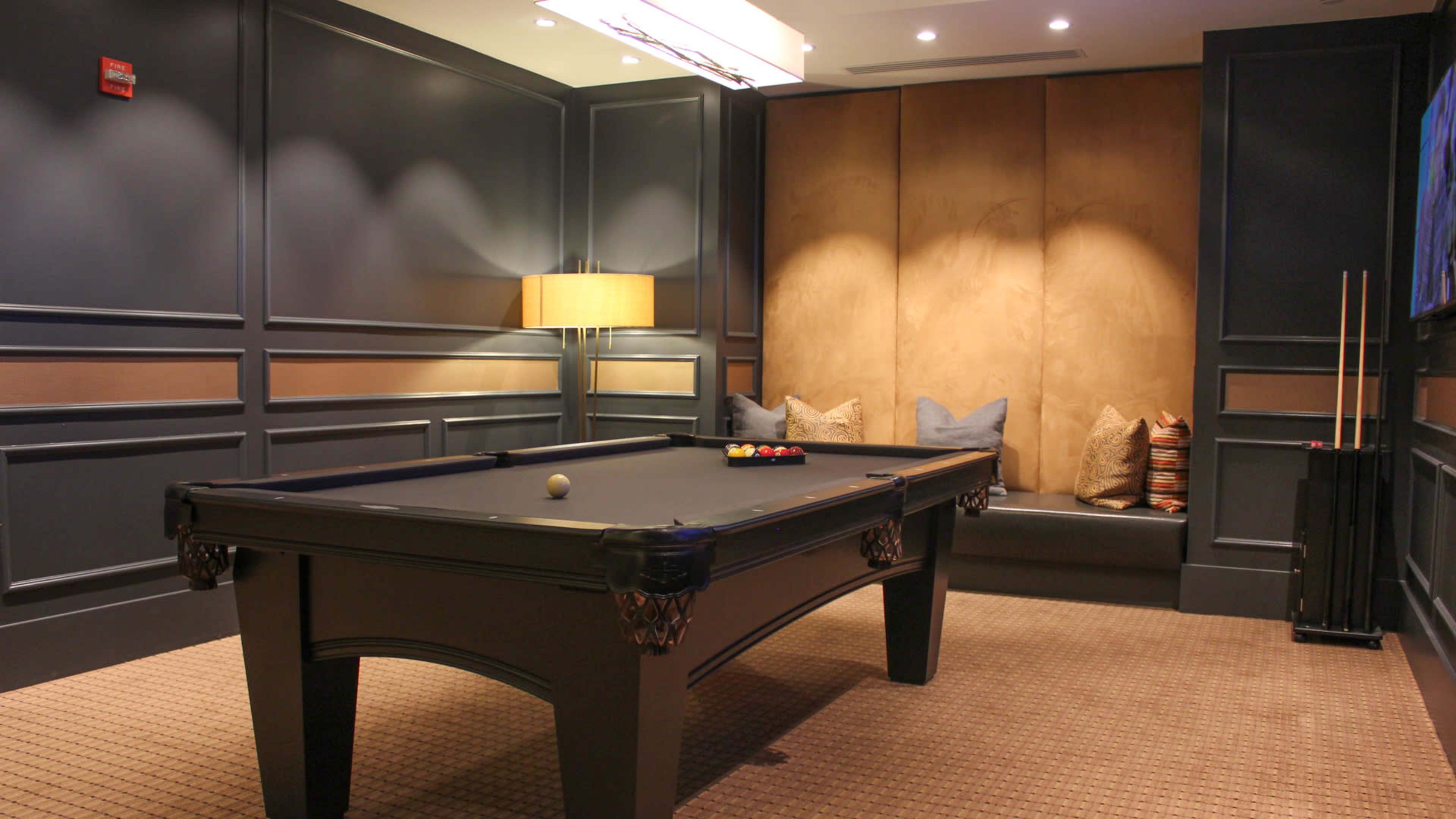 A billiards table is positioned in a well-lit room featuring a lamp, decorative cushions, and a textured carpet.