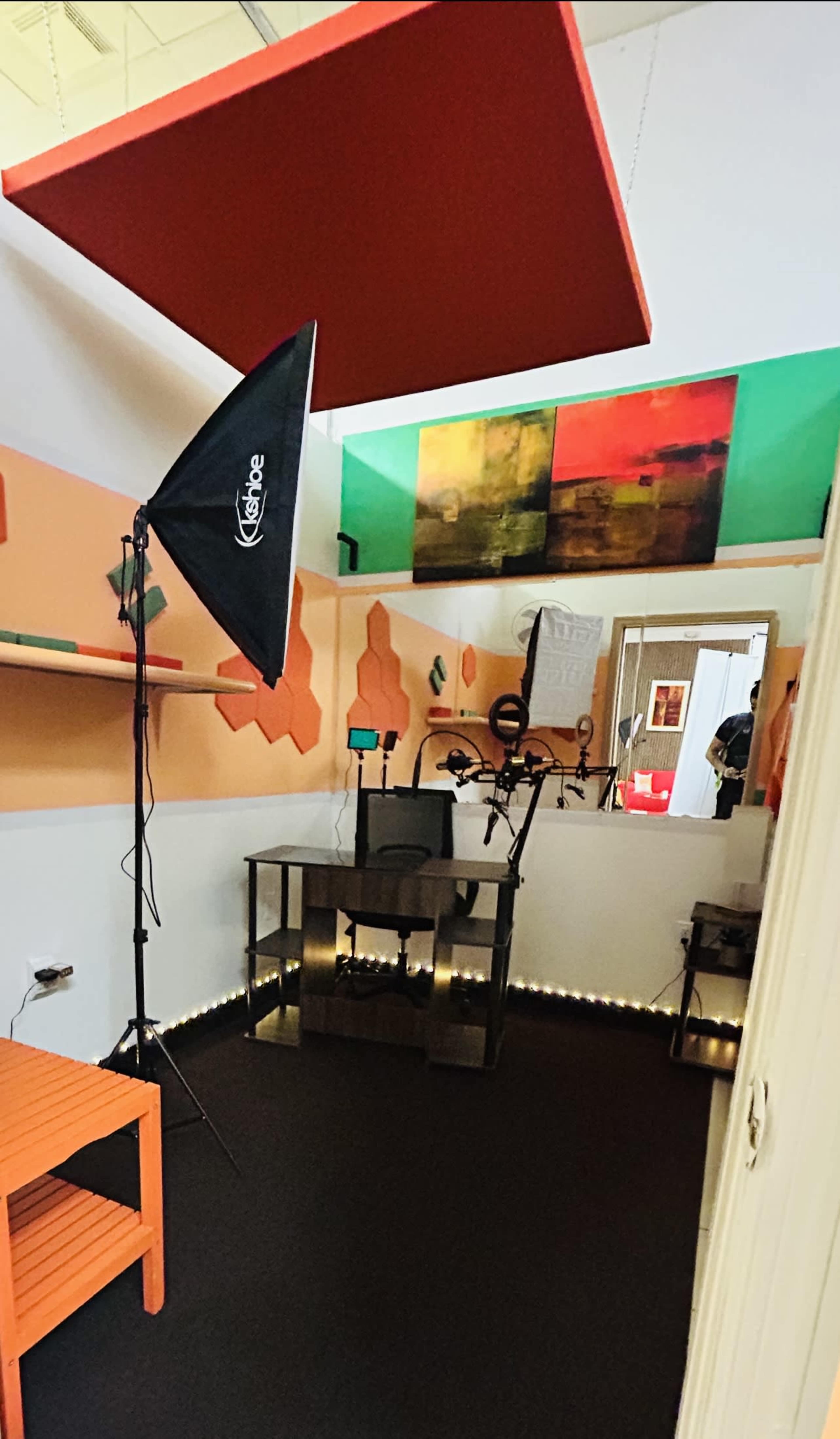 A small room equipped with a lighting setup, a mirror, and a black table under decorative wall art and orange accents.