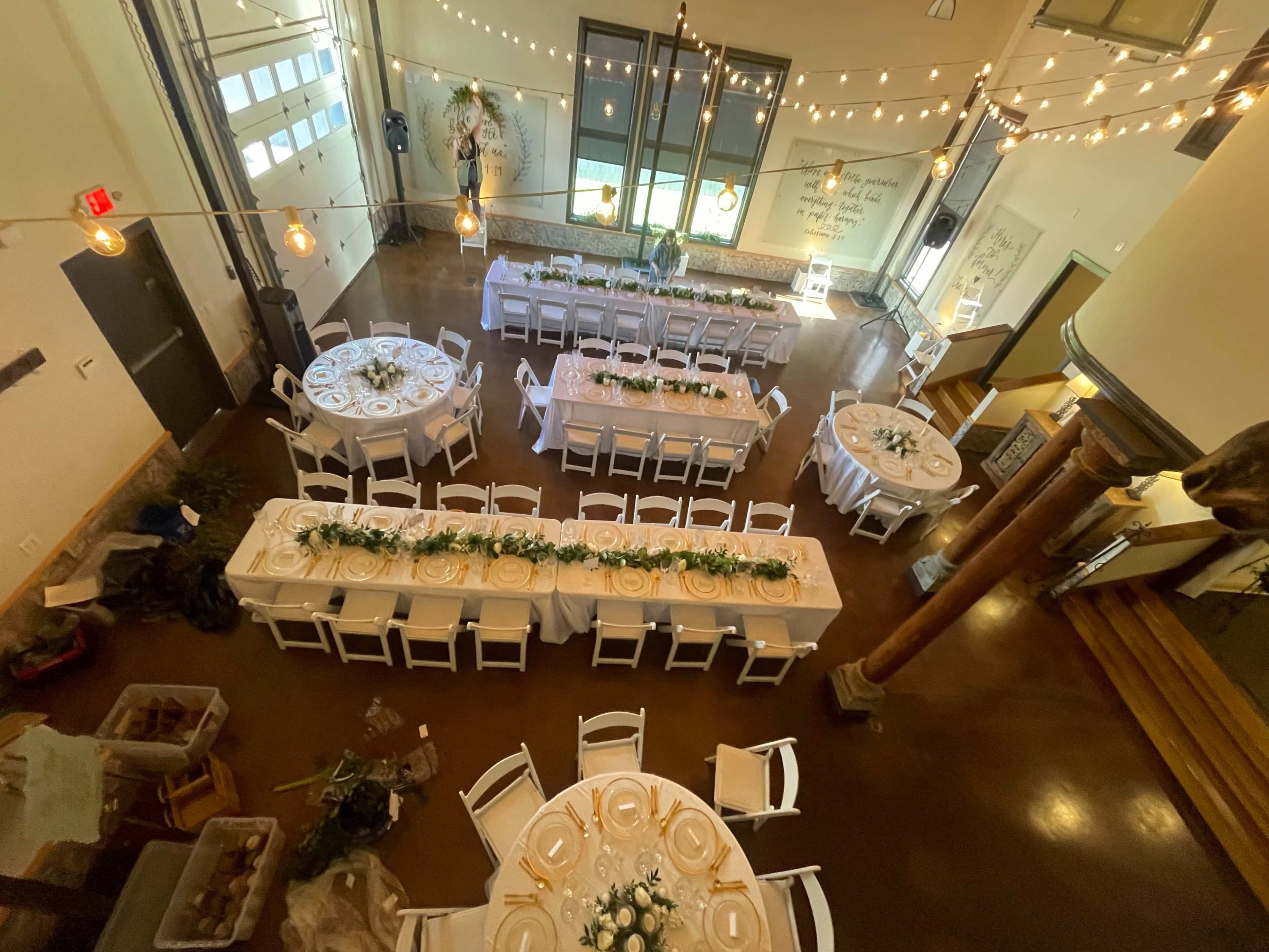 A spacious event hall arranged with multiple circular and rectangular tables decorated with white tablecloths and floral centerpieces, illuminated by string lights.