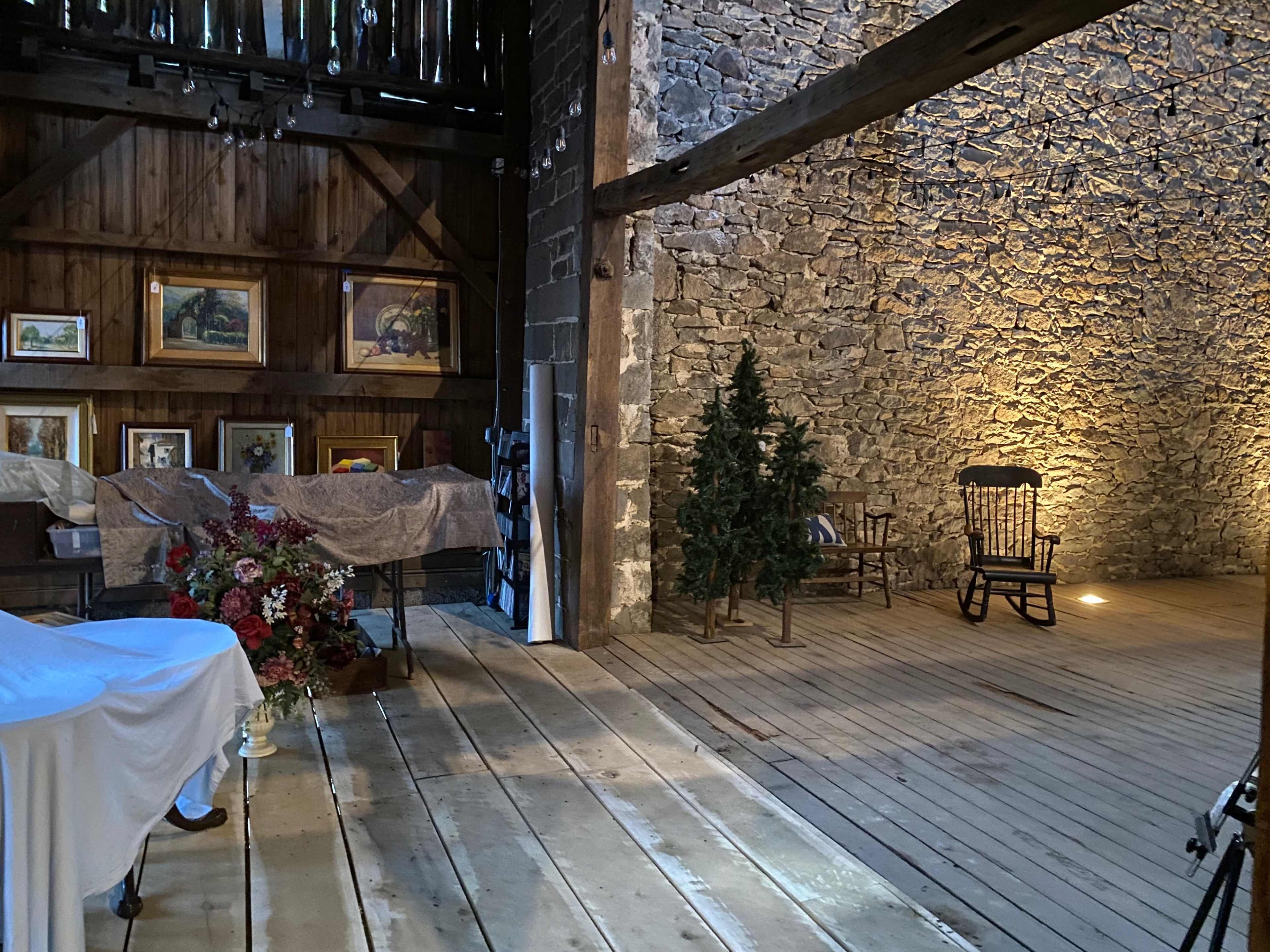 The interior of a rustic barn features stone walls, wooden beams, a wooden floor, and displays of framed paintings alongside a floral arrangement and a rocking chair.
