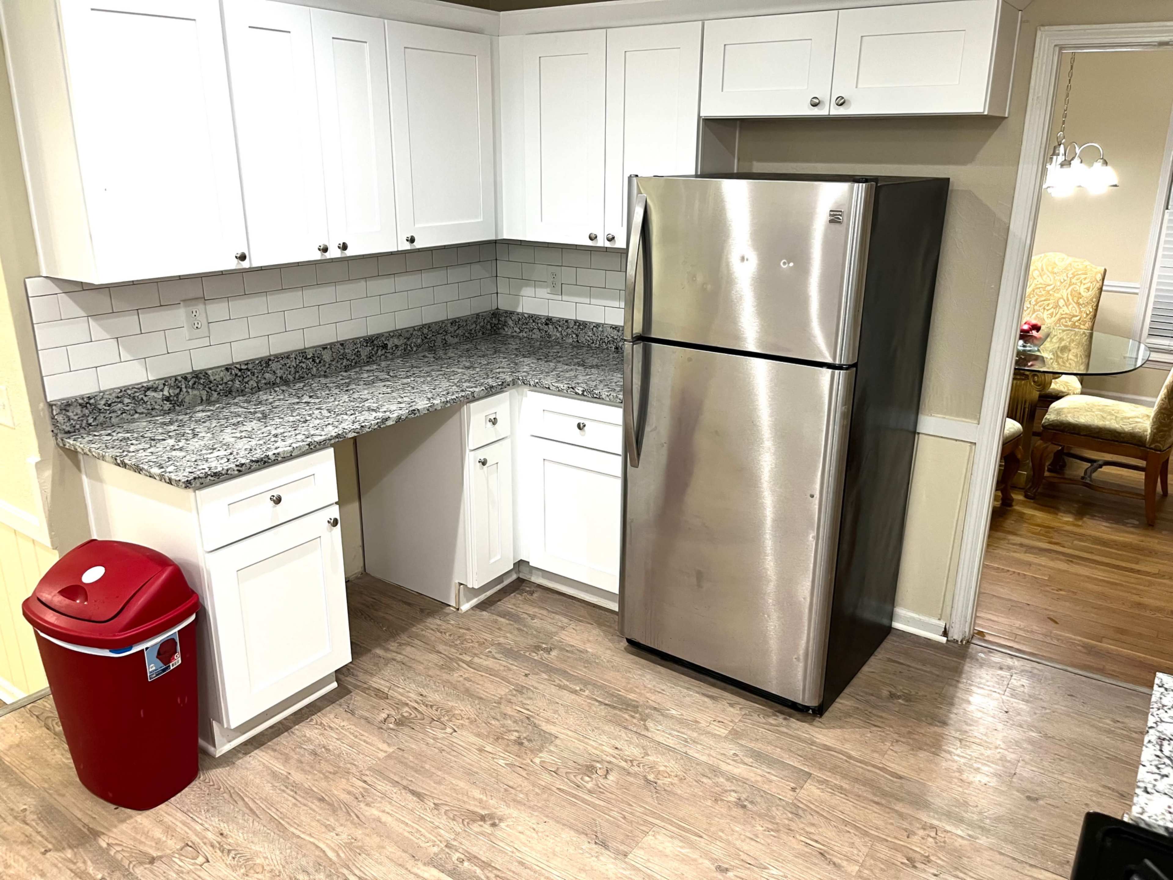 The image shows a kitchen with white cabinets, a stainless steel refrigerator, and a red trash can next to a granite countertop.