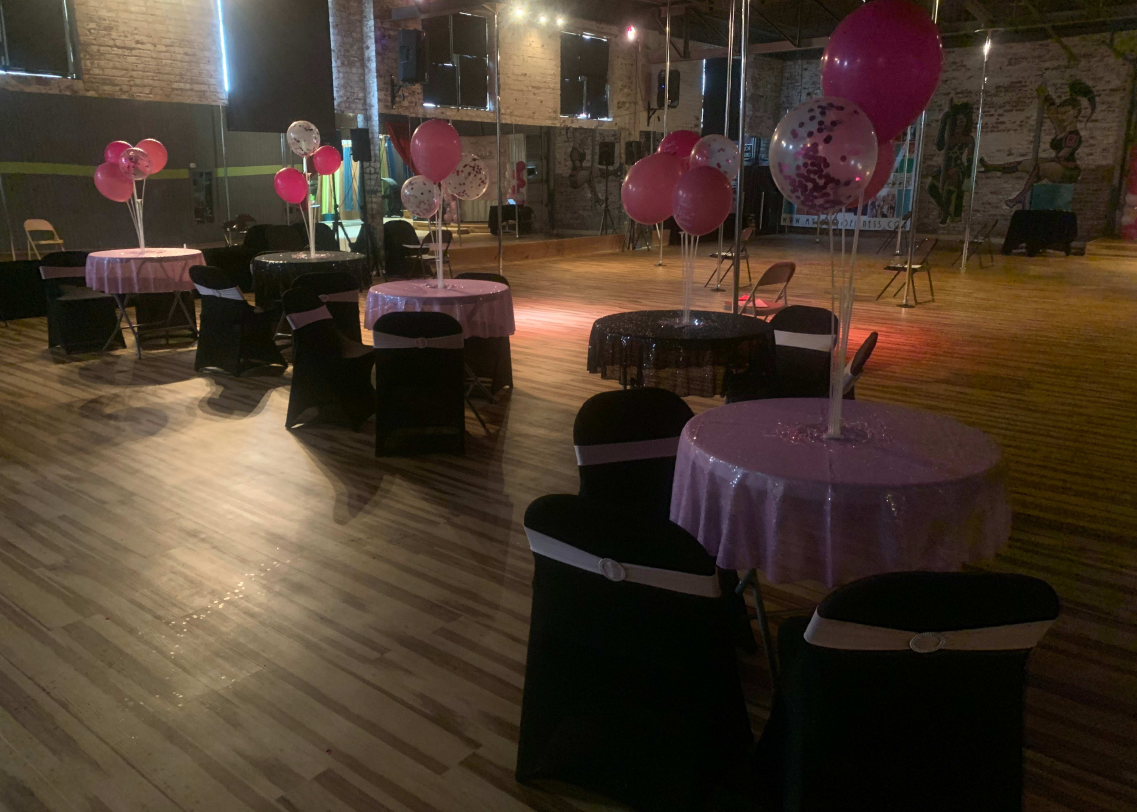 A spacious event venue with tables covered in pink tablecloths and black chairs, decorated with balloons and set up for a gathering.