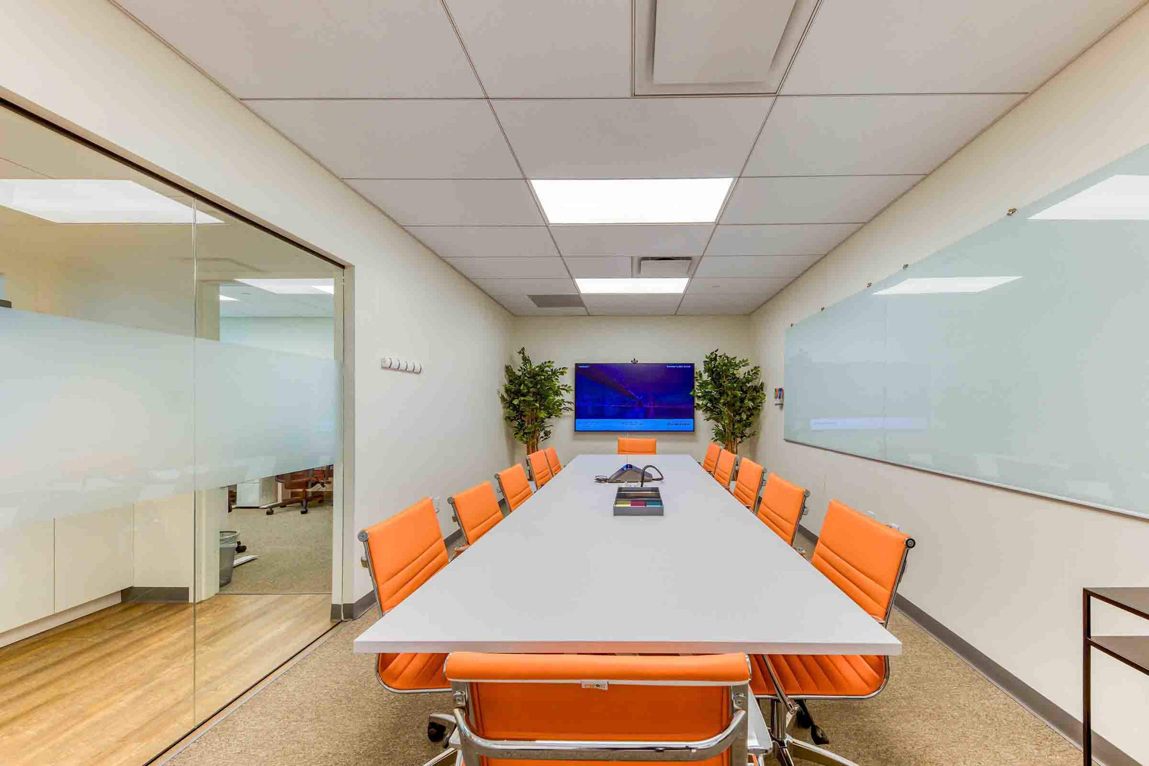 A modern conference room features a long table with orange chairs, a large screen on the wall, and whiteboards on either side.