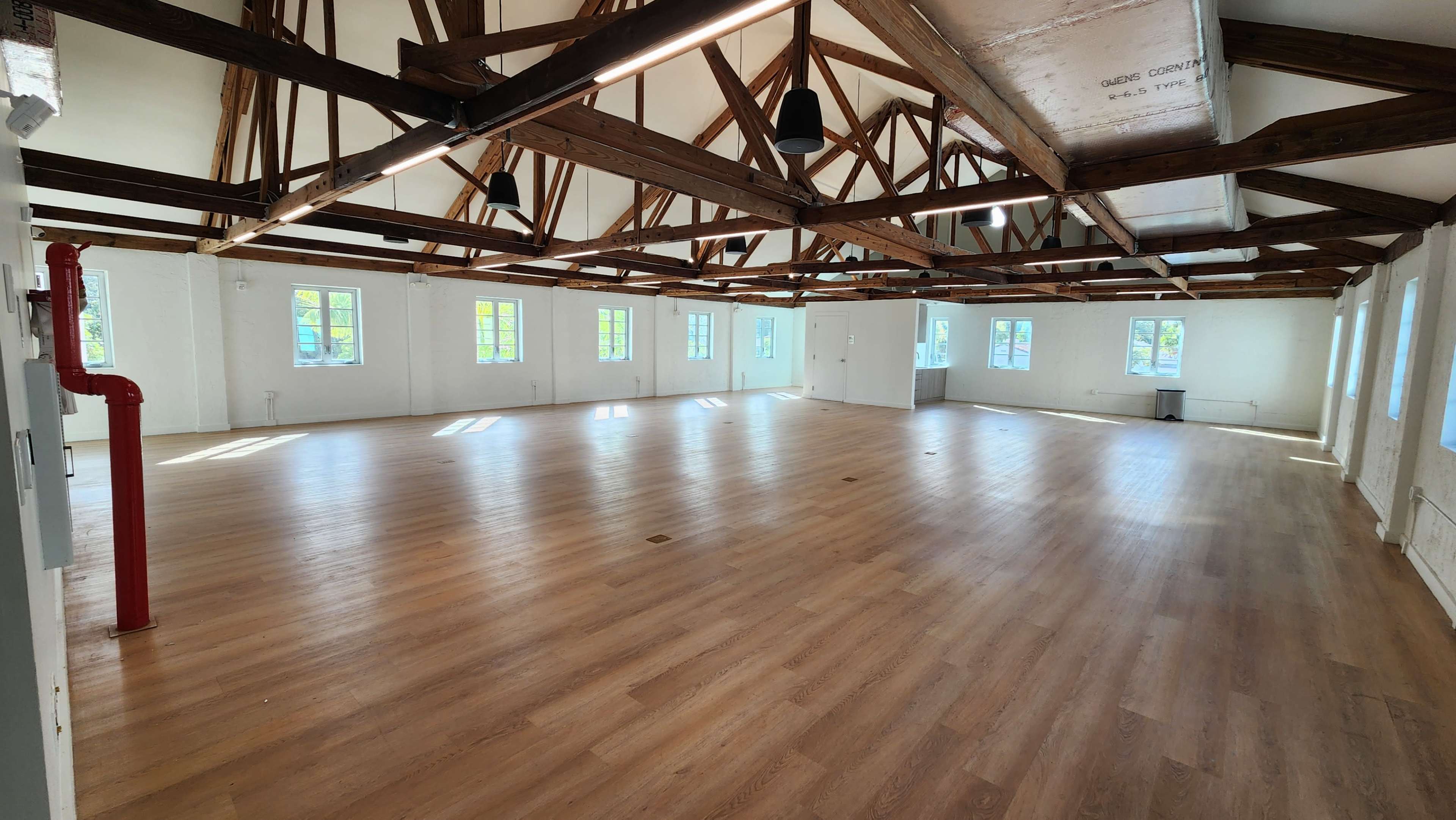 The image shows a spacious, empty room with wooden flooring and a high ceiling supported by exposed beams, featuring numerous windows that allow natural light to enter.