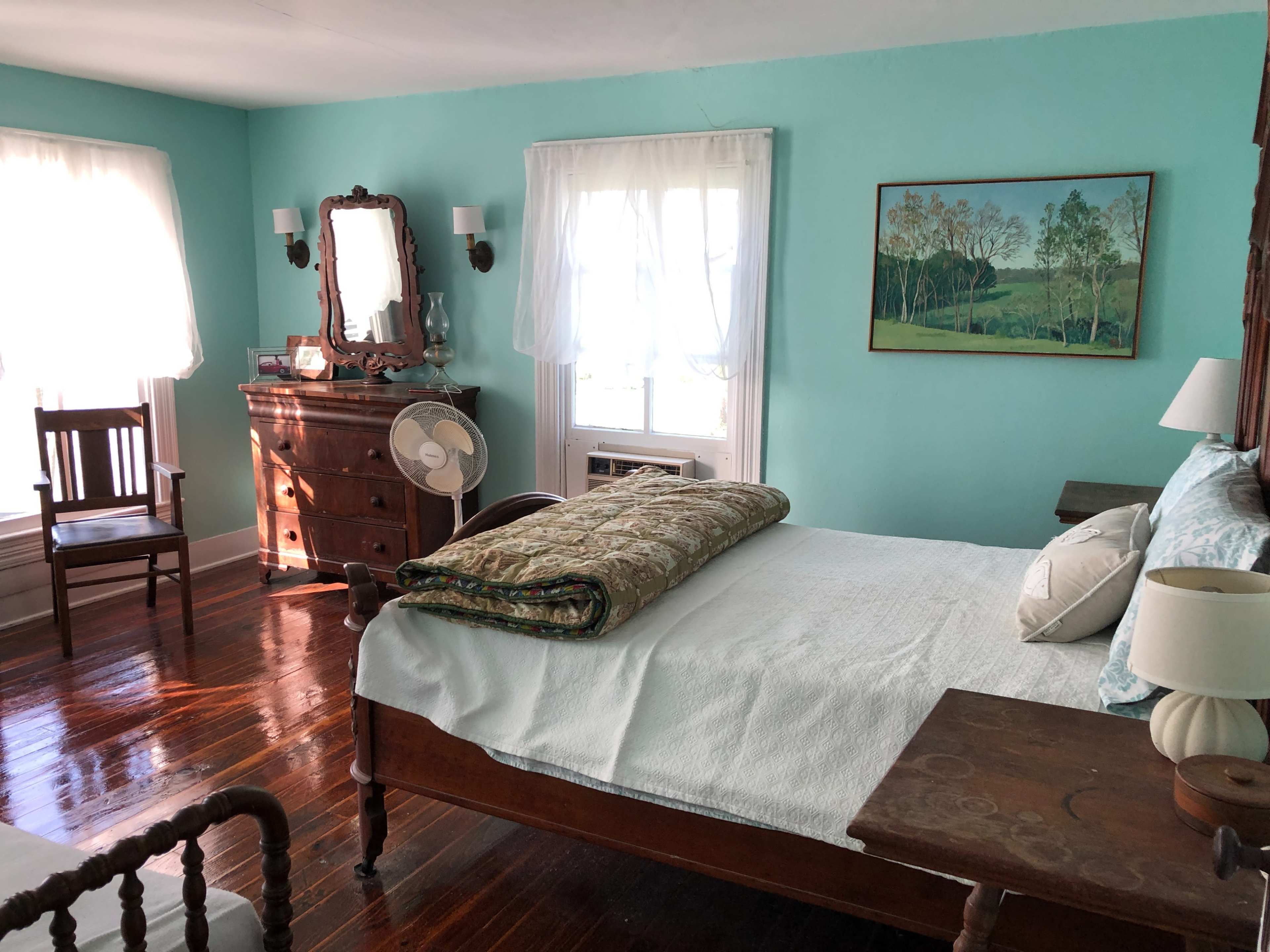 A well-lit bedroom featuring a wooden bed, a fan, a dresser, a chair, and a landscape painting on the wall.