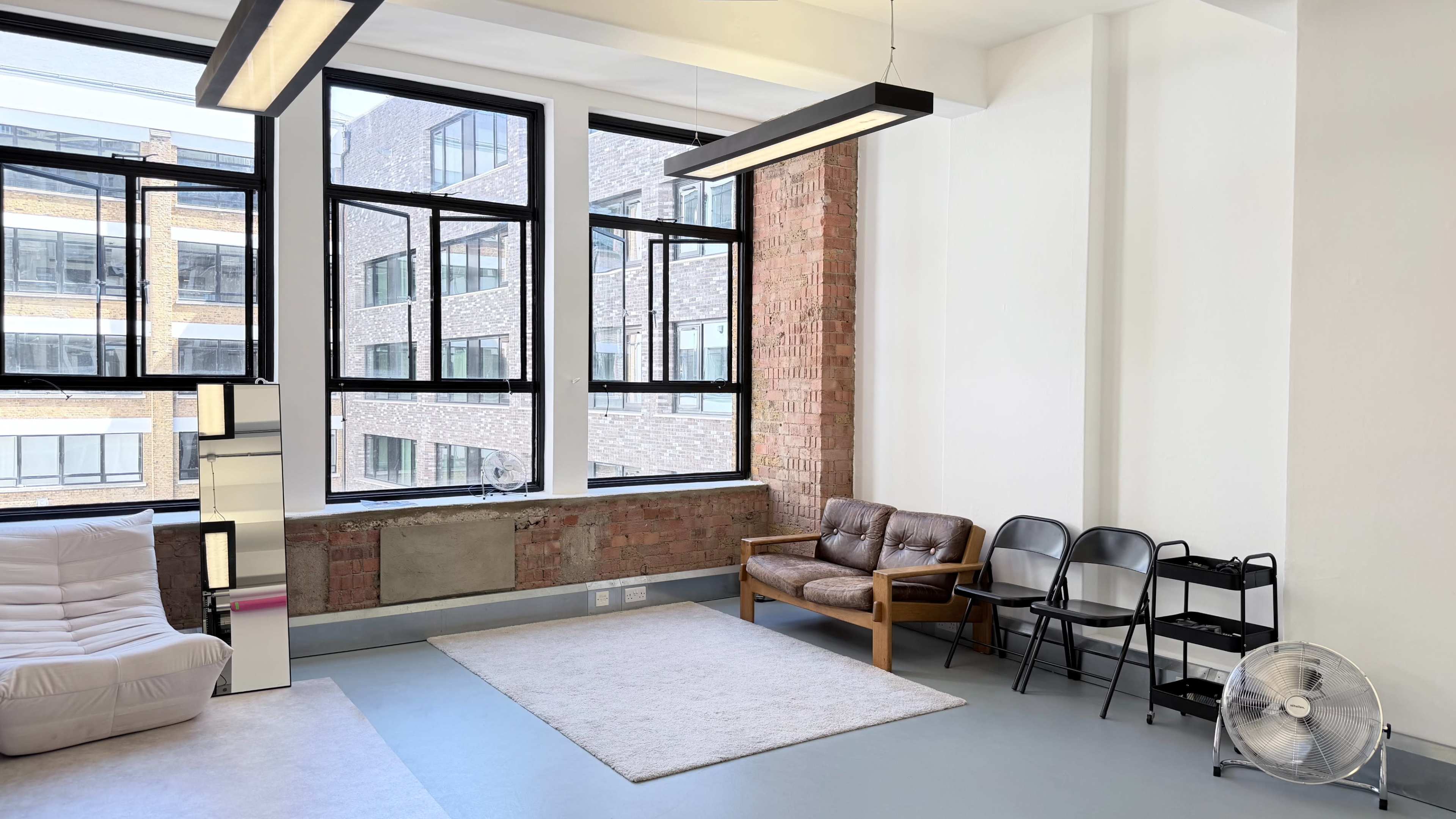 The image shows a bright room with a large window, a white couch, a brown armchair, black folding chairs, and a fan in the corner.