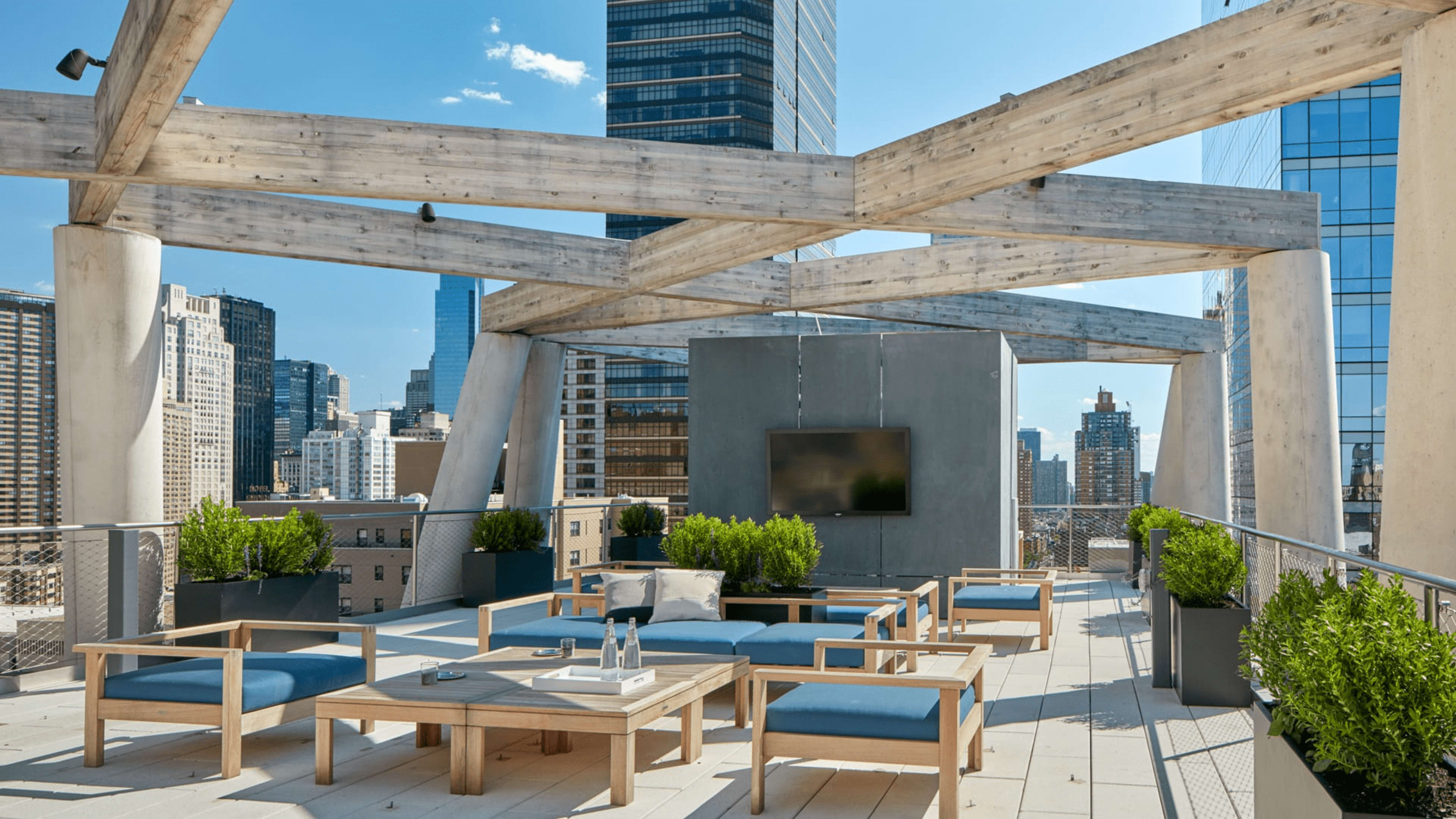 The rooftop terrace features wooden beams, modern seating, and planters, overlooking a city skyline with tall buildings.