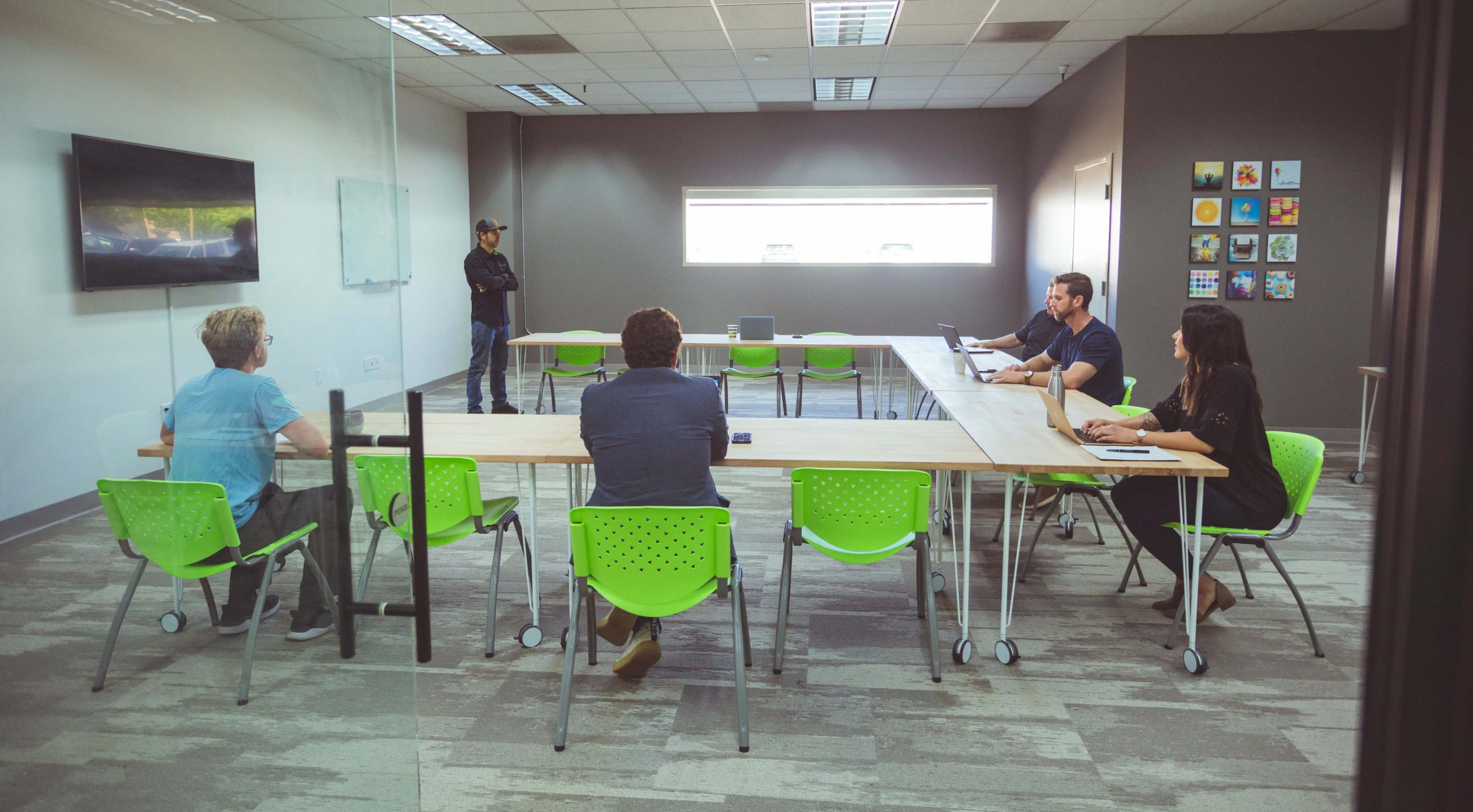 A group of six people sit around a long table in a modern conference room, with green chairs and a large screen in the background.