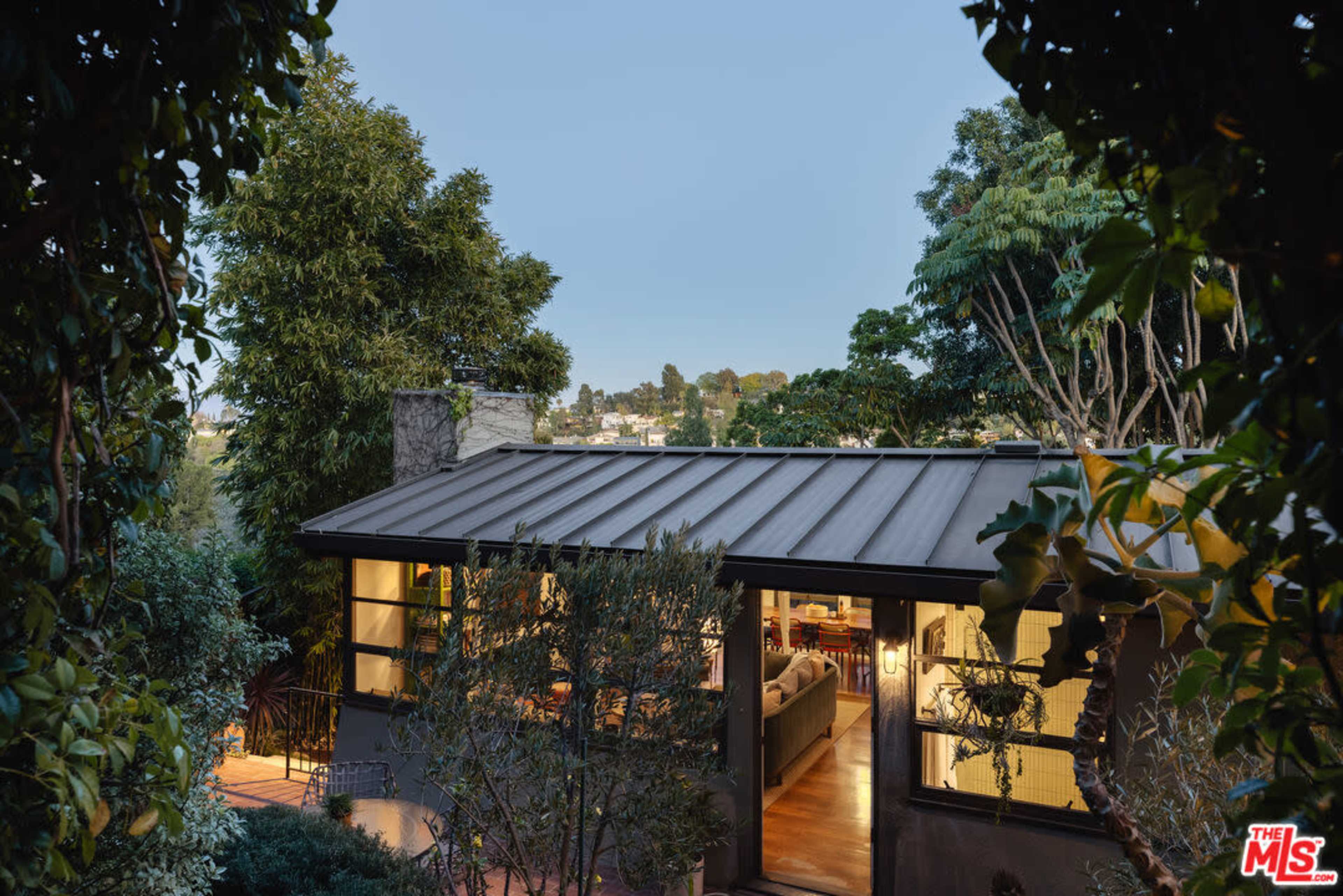 A modern house with a metal roof is nestled among trees, with soft light glowing from its interior.