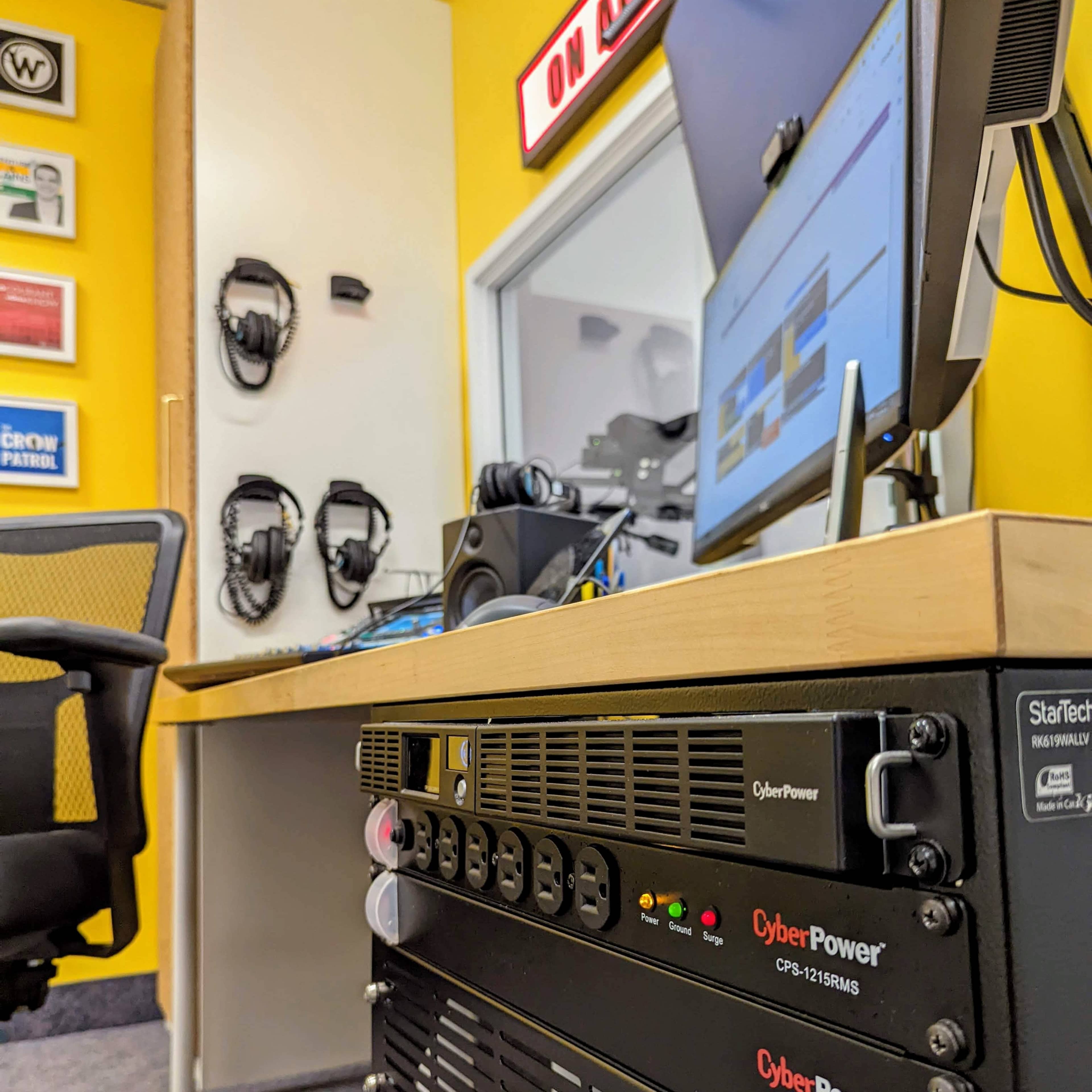 A work desk with a CyberPower backup power supply, computer monitors, and headphones mounted on the wall, set against a yellow background.