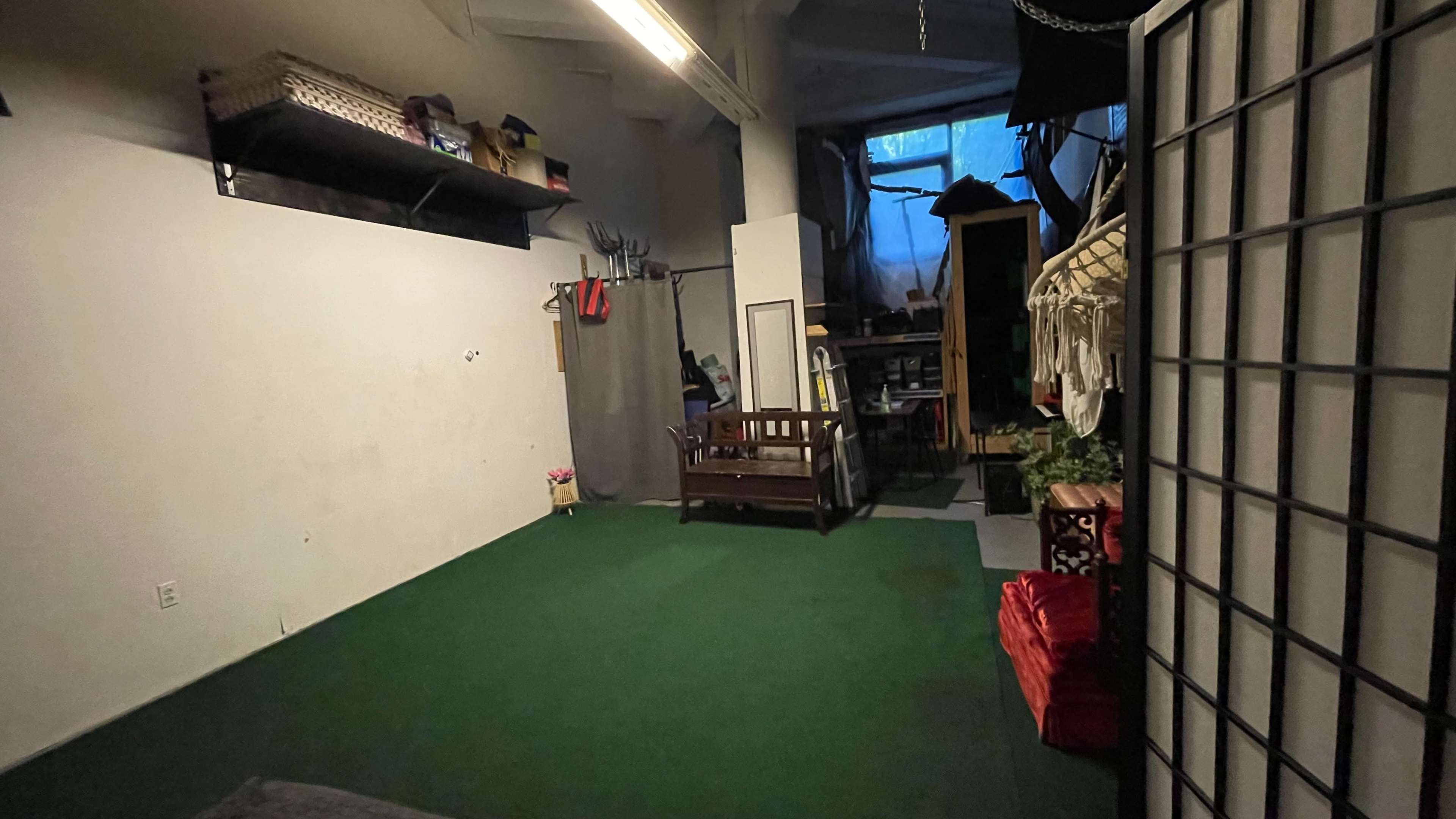 The image shows a spacious room with artificial green carpet, a bench, and various storage items along the walls.