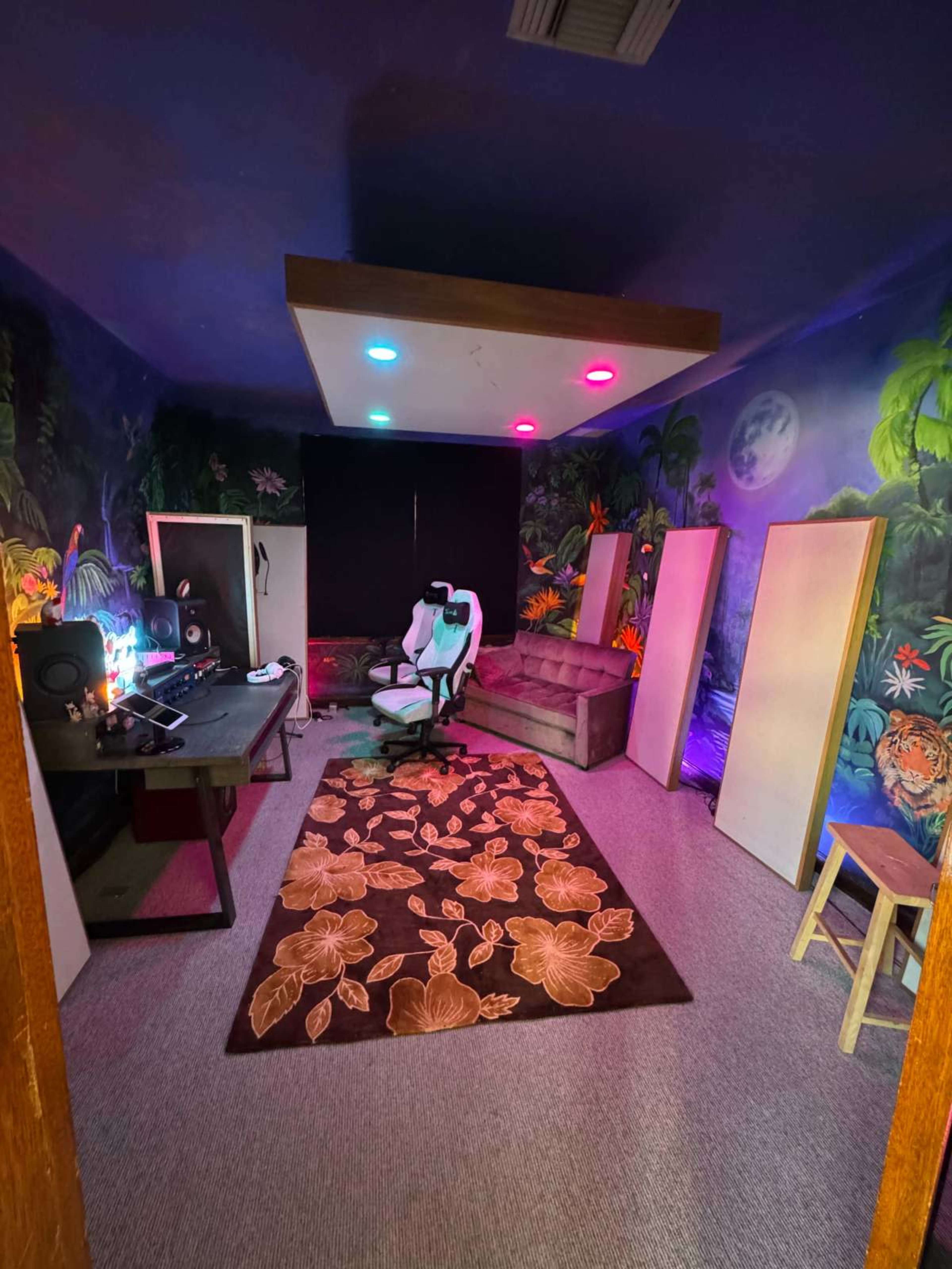 The image shows a colorful, mural-decorated room featuring a gaming chair, a desk with equipment, a sofa, and several blank panels arranged around a patterned rug.