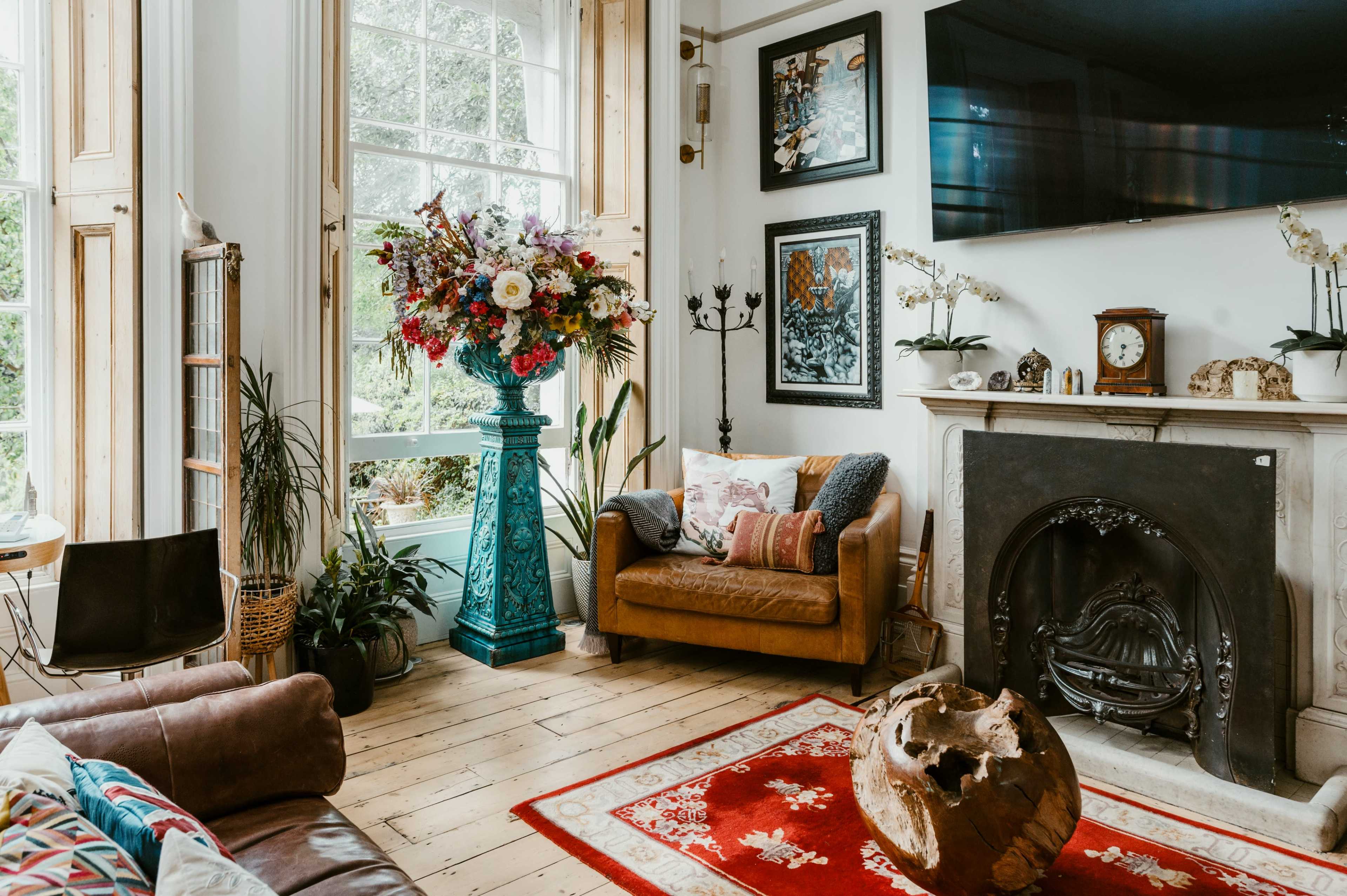 The room features a vintage sofa, a large floral arrangement, a fireplace with ornate detailing, and an assortment of plants and artwork on the walls.