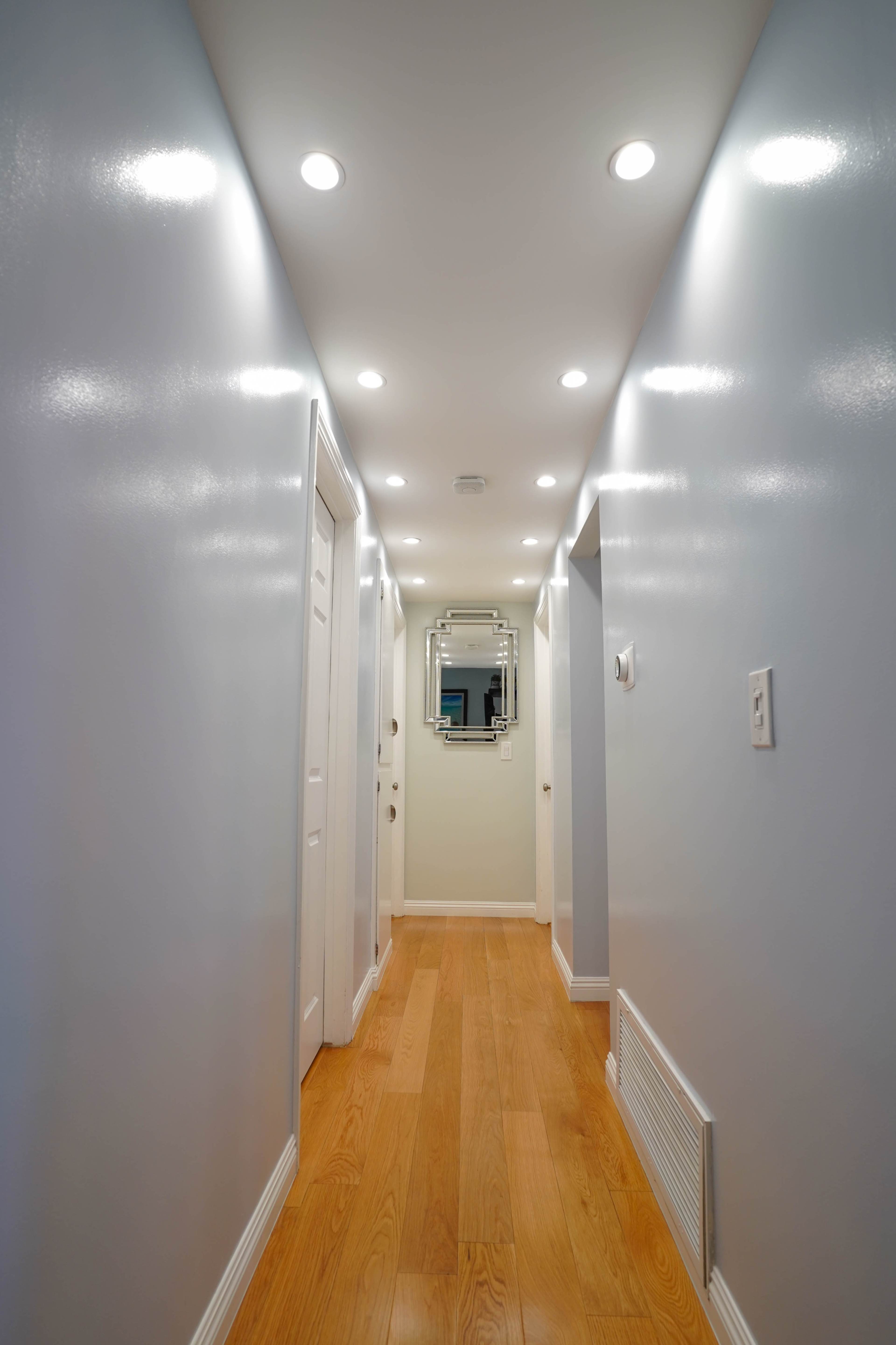 A well-lit hallway with white walls, a wooden floor, and a mirrored cabinet at the end.