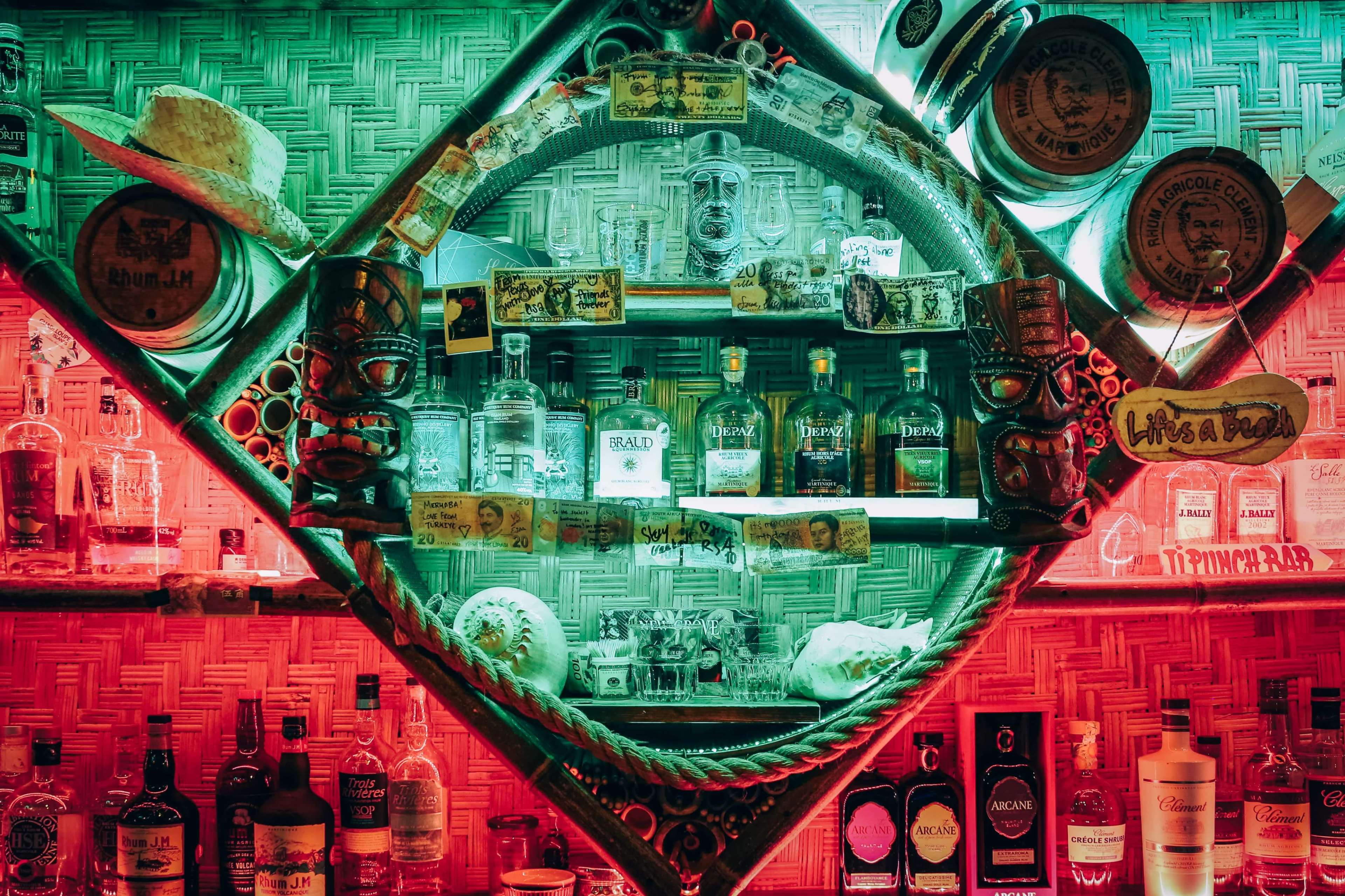 The image shows a bar shelf filled with various bottles of alcohol, framed by a decorative circular arrangement with hats and memorabilia in green and red lighting.