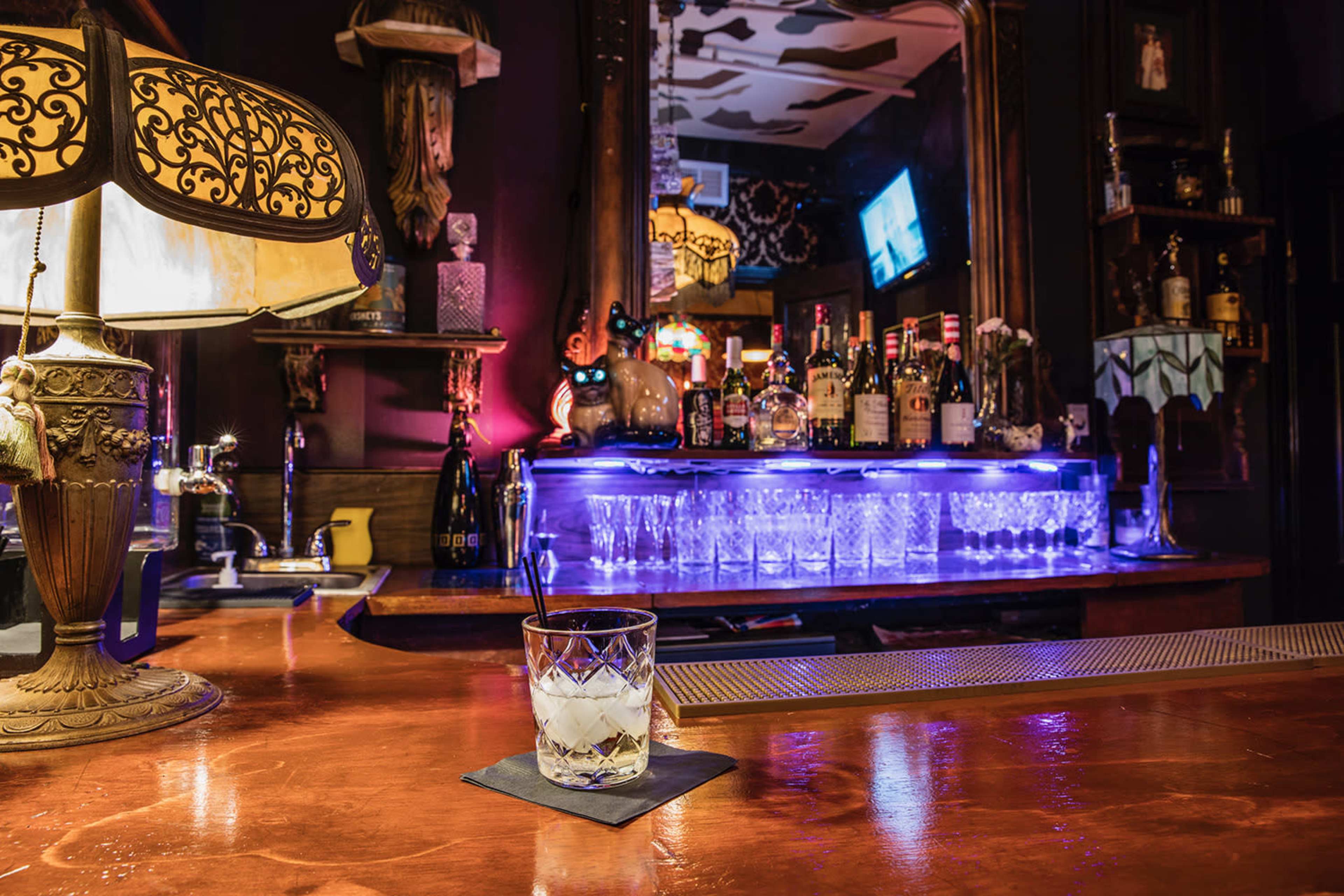 A well-lit bar scene features a glass of cocktail on the counter in front of a backlit shelf stocked with various liquor bottles.