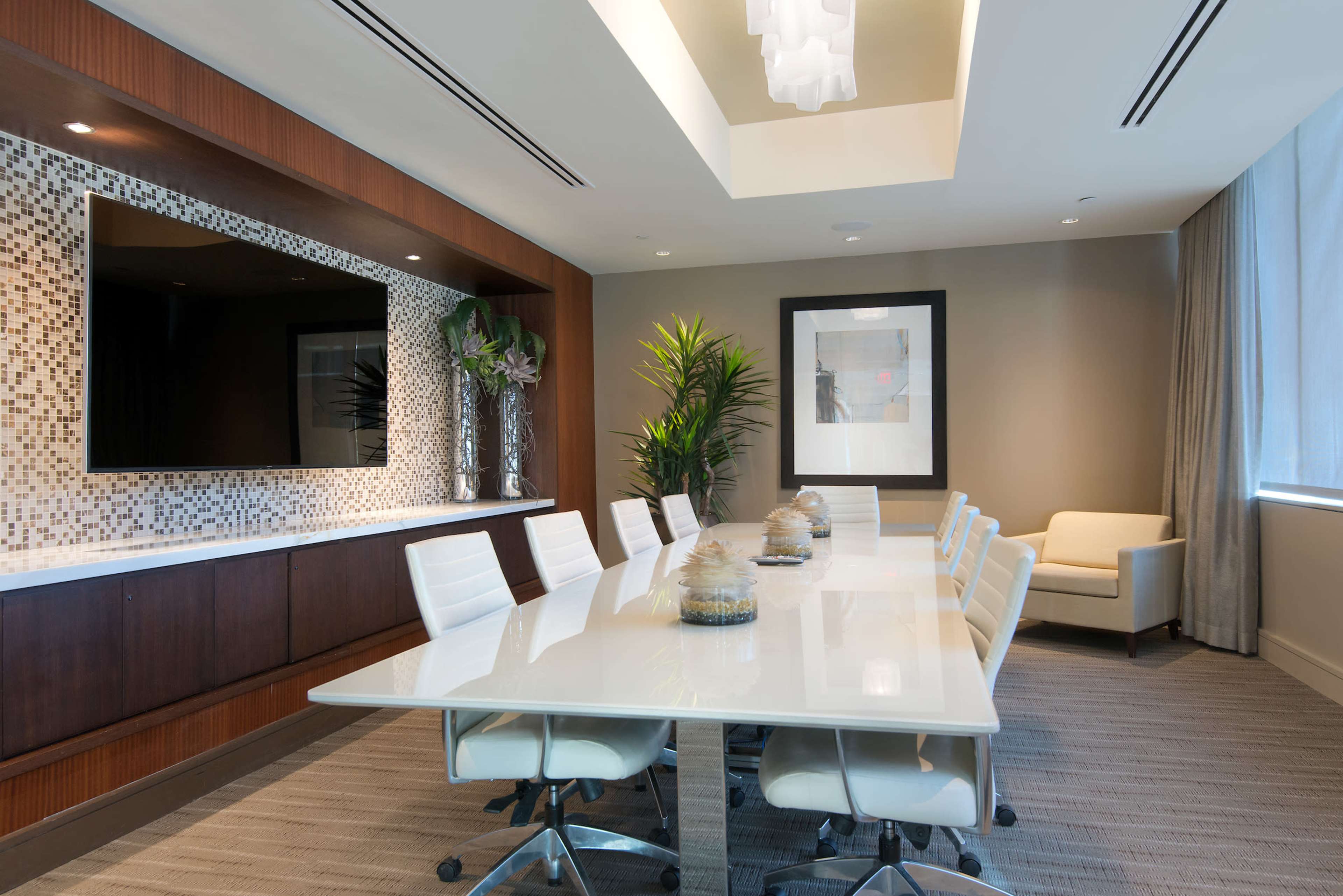 A modern conference room features a long white table surrounded by ergonomic chairs, with a decorative wall, large television, and a window providing natural light.